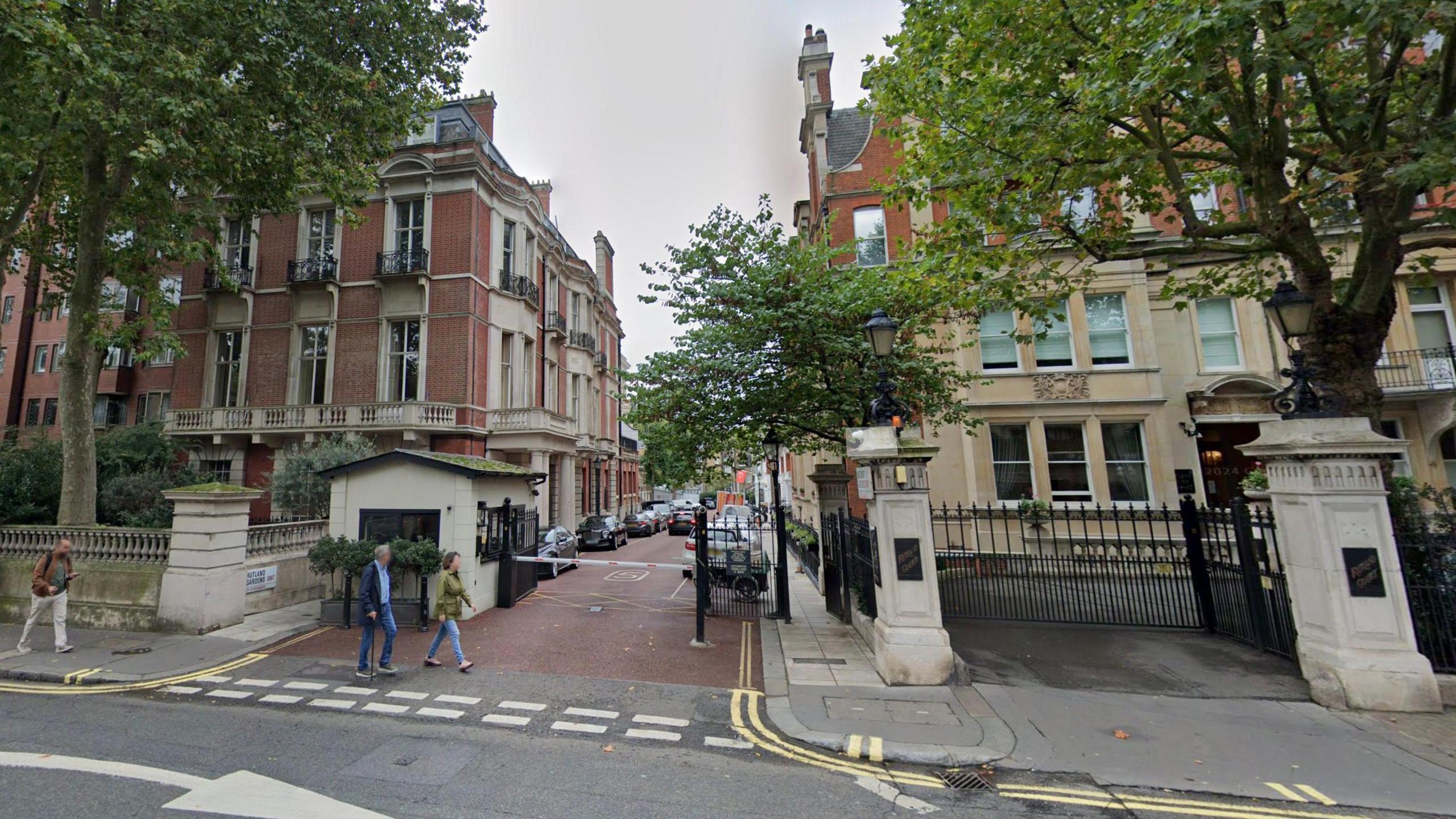 Streetview of Rutland Gardens shows pedestrians walking past tree-lined street with pastel coloured buildings and a barrier across the entrance