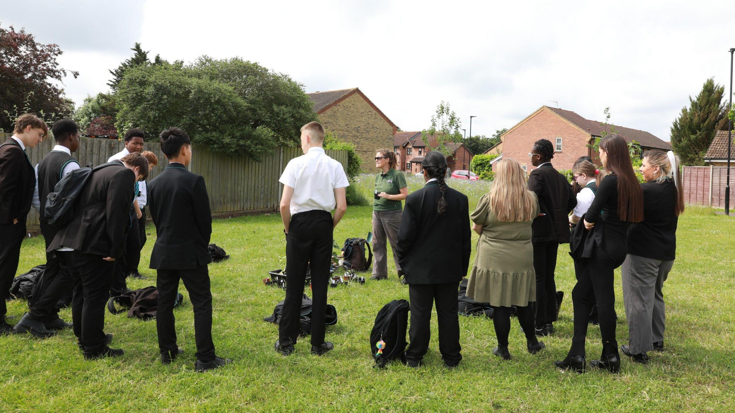 A group of students wearing a black uniform with a white shirt, from Rivers Academy West London, are stood facing a lady wearing a green T-shirt. They are on a patch of grass outside, tasked with turning wasteland into an orchard.