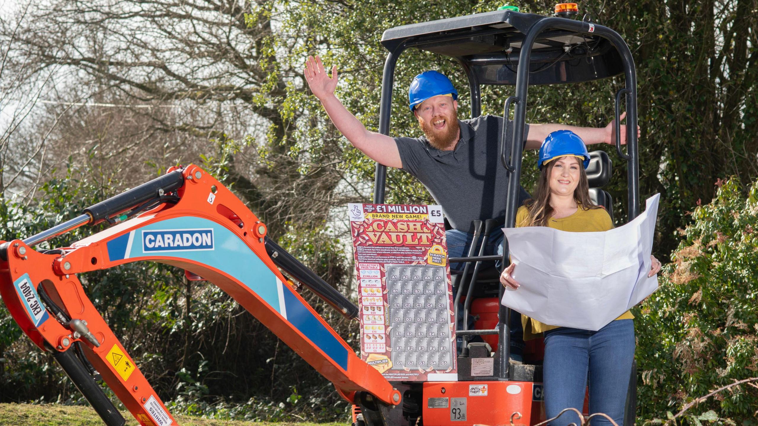 Zoe and Craig wearing hard hats that are blue, Craig is sat in an orange digger and Zoe is holding blue prints for plans.