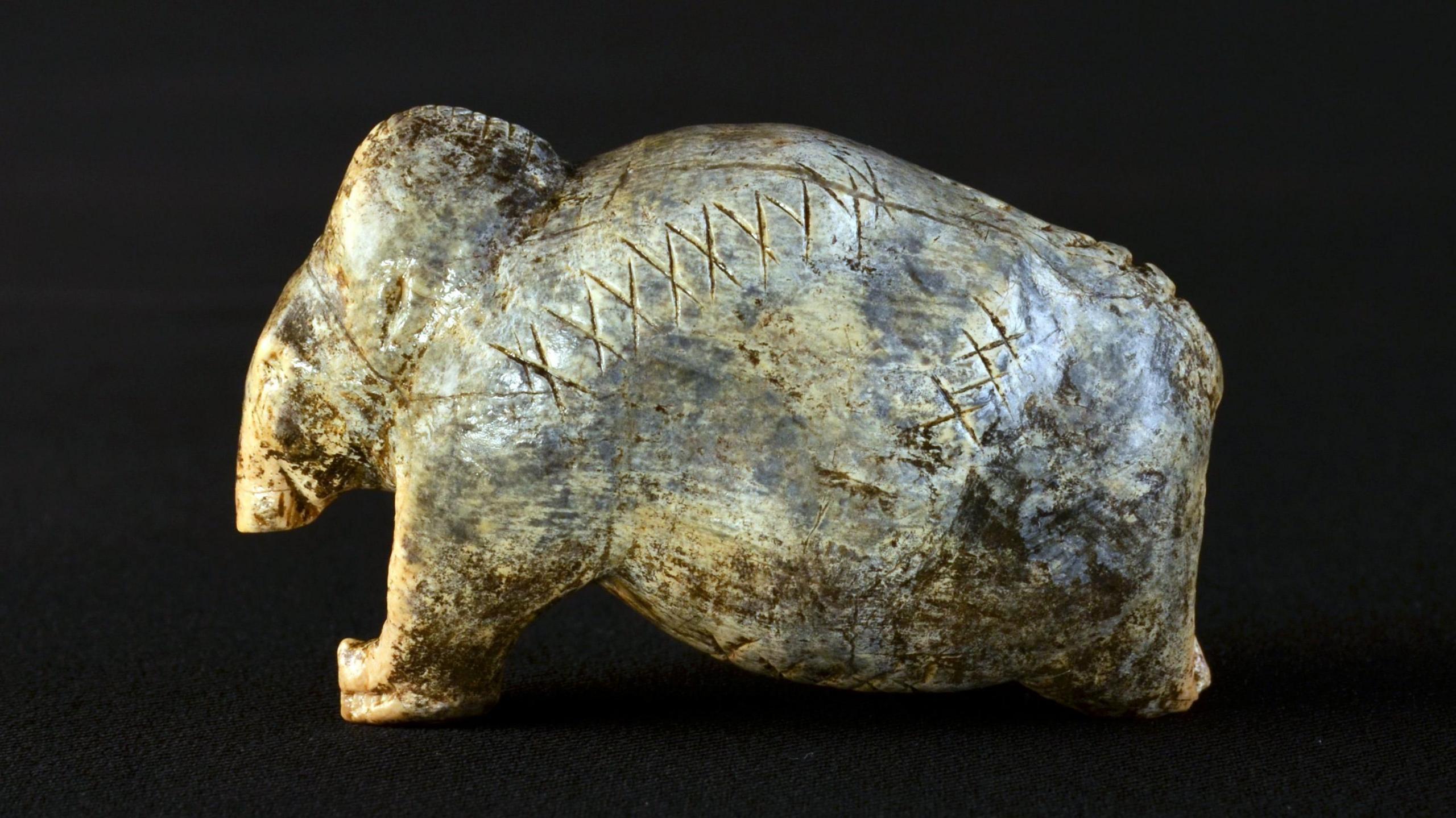A figurine shaped like a mammoth. It is ivory colour with dark grey patches, and has deeply-engraved lines of crosses and dots
