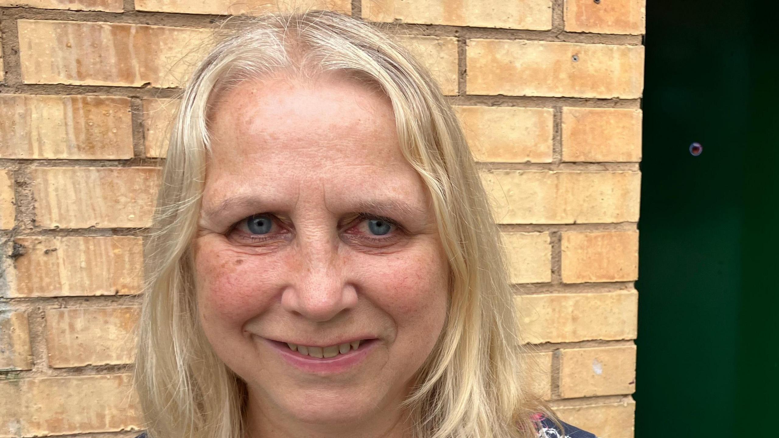 Sheena O'Sullivan is looking slightly to the right of the camera. She has blonde hair, is smiling, and is standing in front of a brick wall