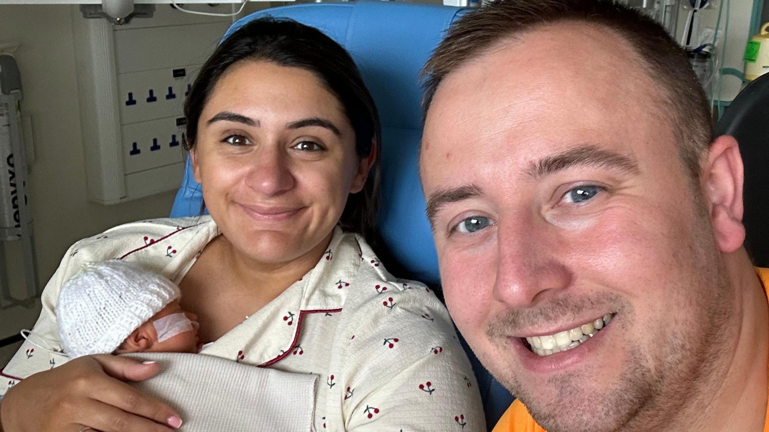 Aisha McCracken in pyjamas in hospital holding her newborn baby who is wearing a white woolly hat and is covered by a blanket. Husband David, wearing an orange T-shirt, is also in the picture. The couple are both smiling at the camera.