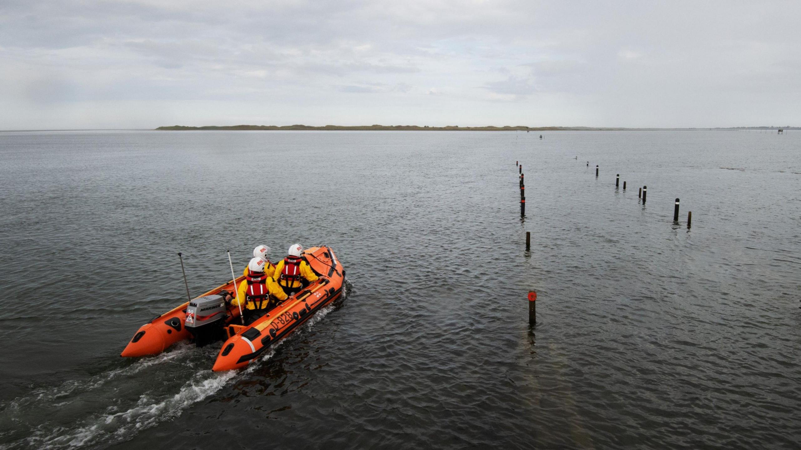 A small inflatable lifeboat with three crew members makes its way across the Holy Island causeway. Land can be seen in the distance.