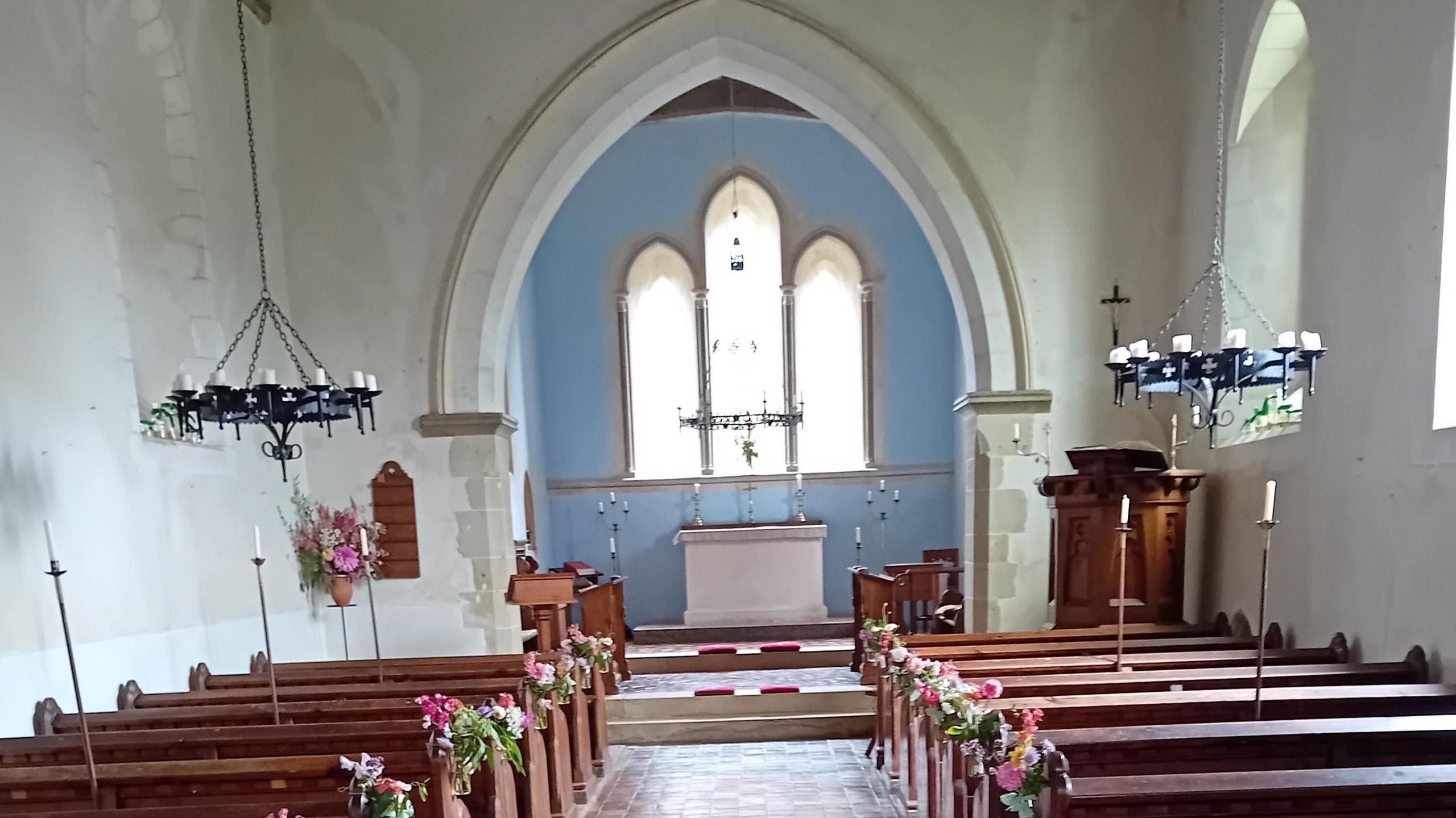 An internal image of St Leonard church in South Stoke, West Sussex. The wall behind the white altar is painted sky blue, while pink flowers can be seen at the end of each row in the church. There are two candle chandeliers above the rows.