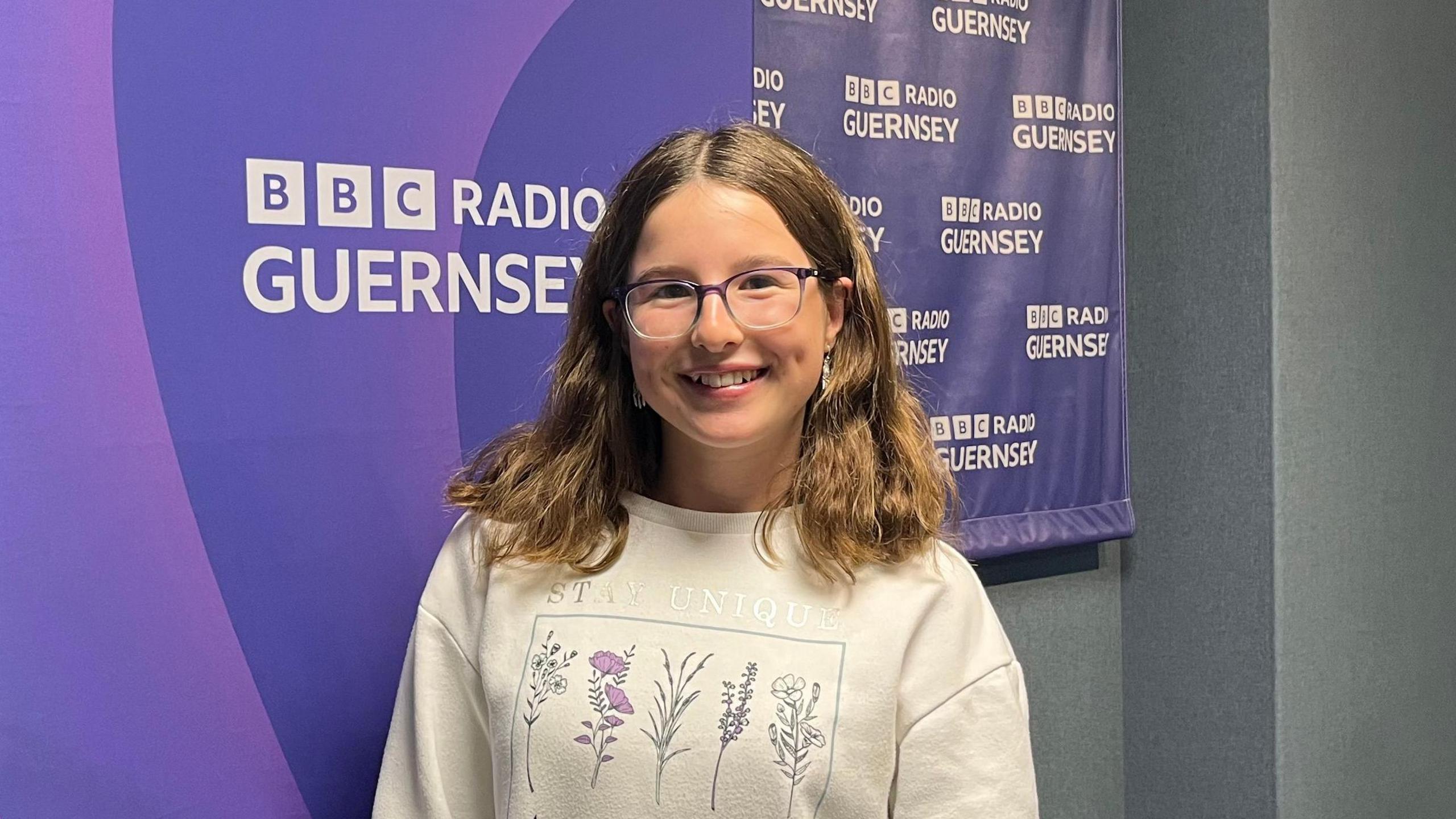 A smiling girl has shoulder length brown hair and is wearing glasses. She is standing in front of a purple banner with the BBC logo and words Radio Guernsey picked out in white.