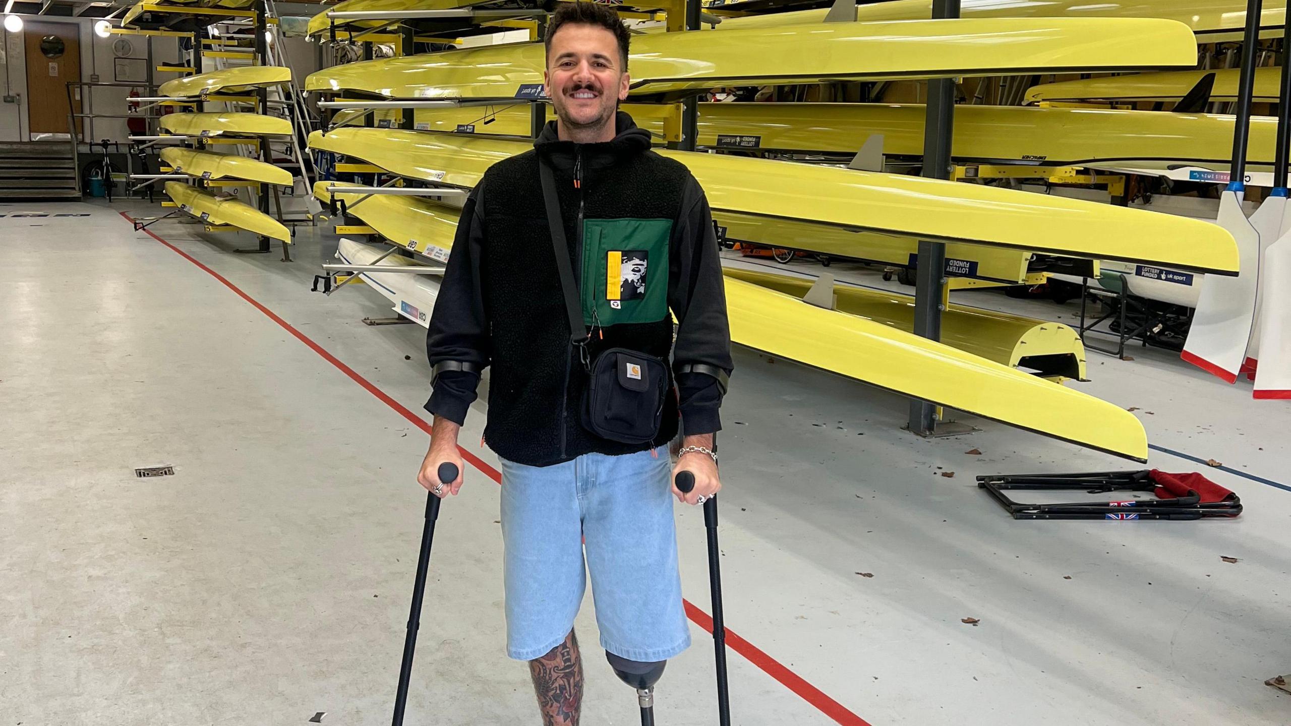 Luke Tarrant, holding two crutches, standing by several rows of yellow rowing boats on racks. He is smiling, wearing denim shorts, a black top, with a bag across him. He he has short dark hair. One leg has lots of tattoos. 