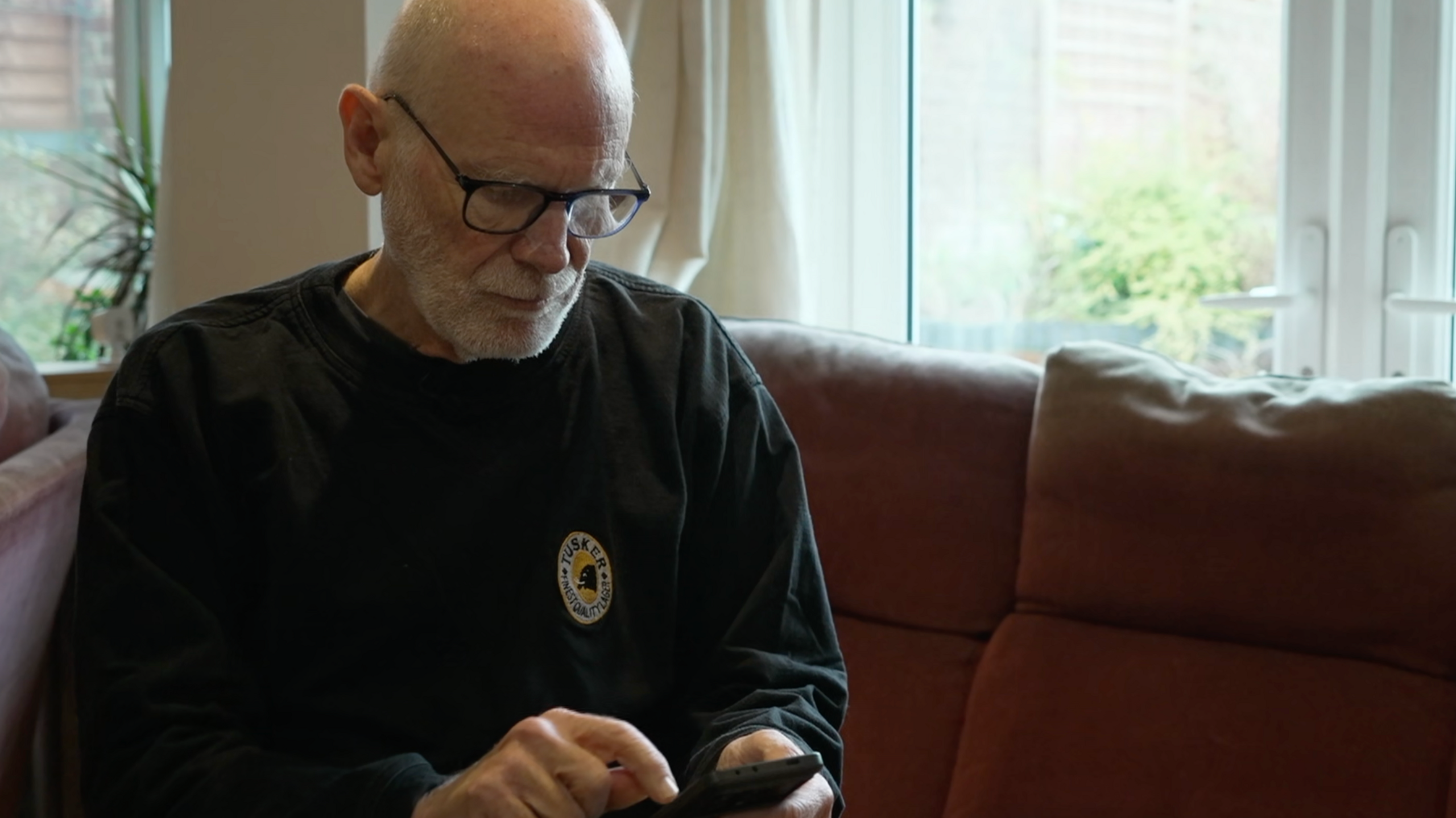 Gideon Rabinowitz wears a dark jumper and sits on his sofa looking down towards his mobile phone