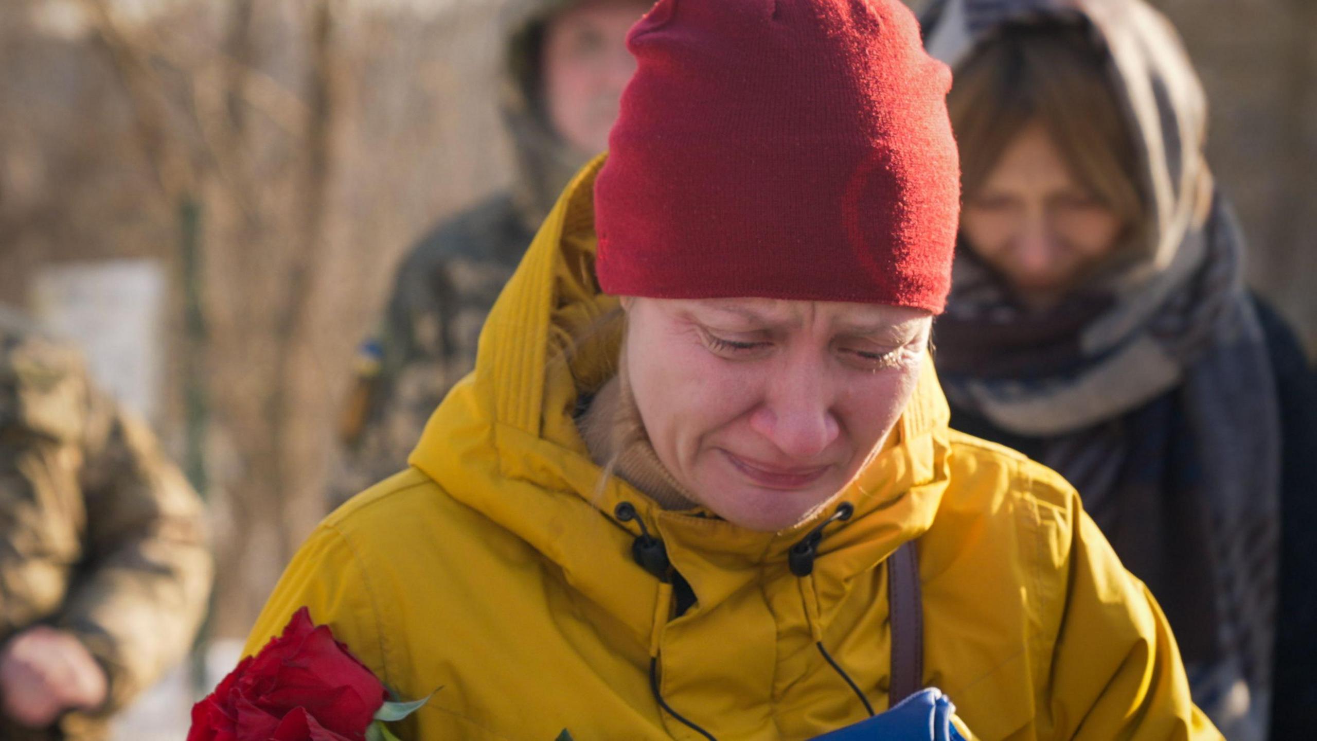 Natalia weeps by the grave of her husband in a snow-covered cemetery in Kyiv. She's dressed in a yellow jacket and a red beanie hat, and is holding a red rose. 