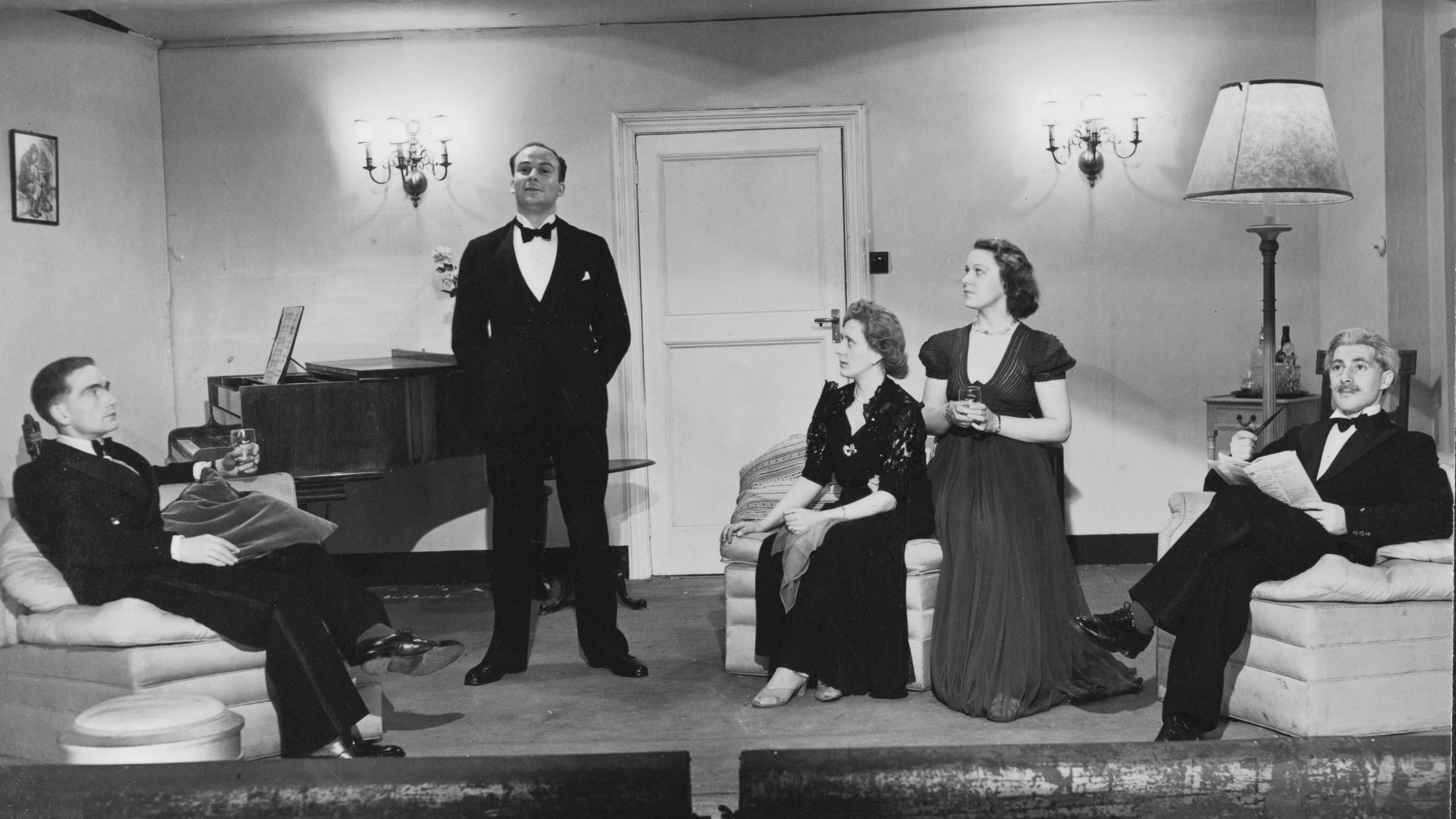 Three men and two women are sitting and standing in a mock front room on stage. The men are in dinner suits and bow ties, and the women have long evening dresses on. There are arm chairs, a grand piano and a large lamp on a side table.