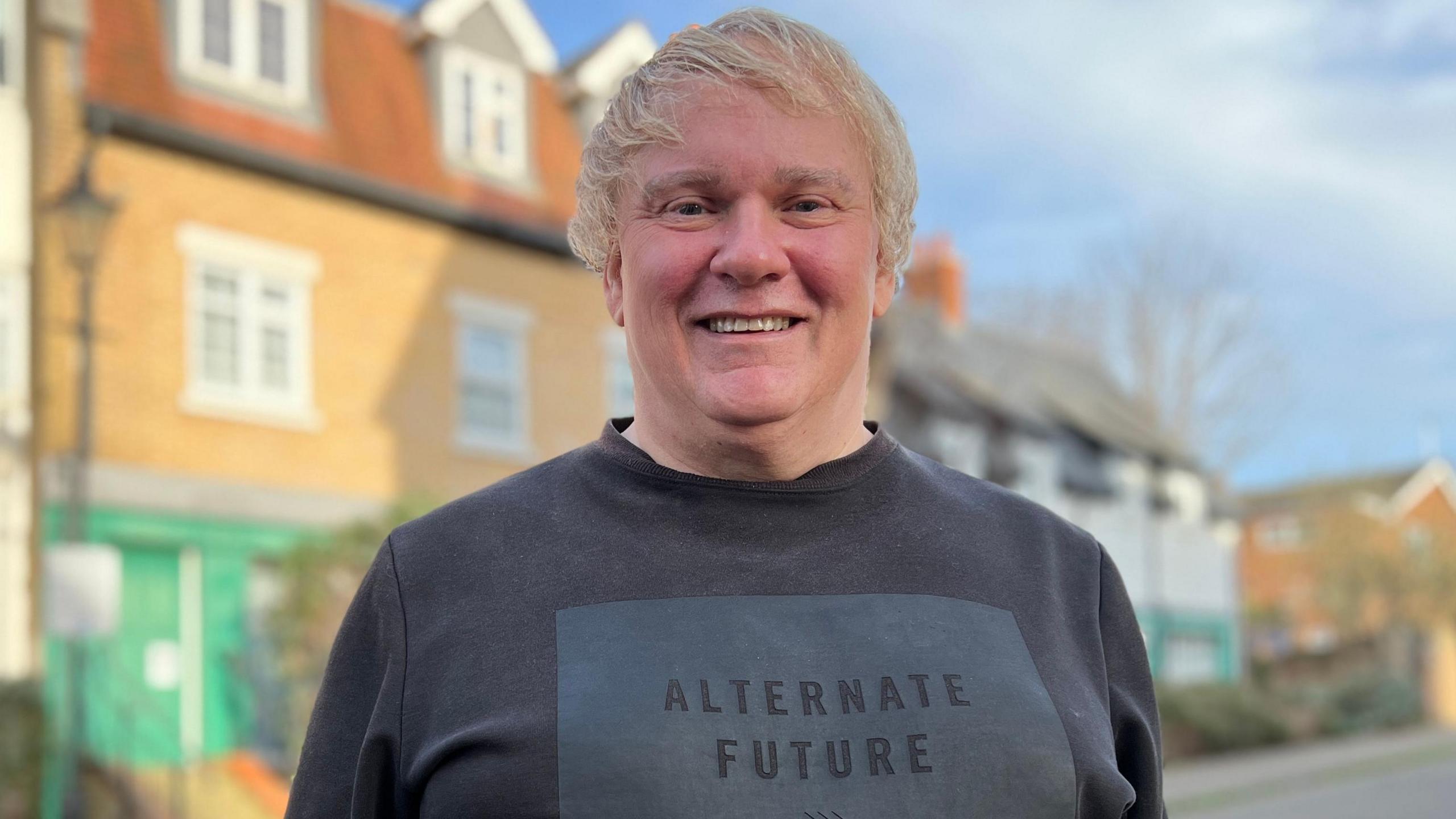 David Davensac, stood wearing a black t-shirt in front of a row of houses with blue skies above.