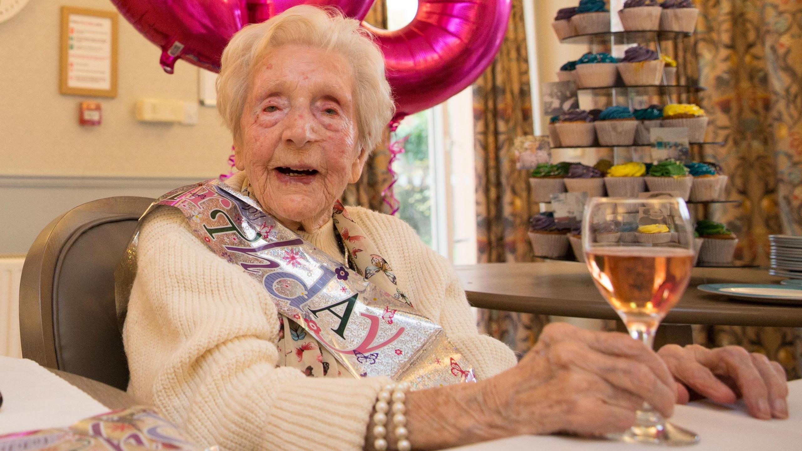 105-year-old Daisy Taylor with a birthday sash. She sits at a table holding a glass.