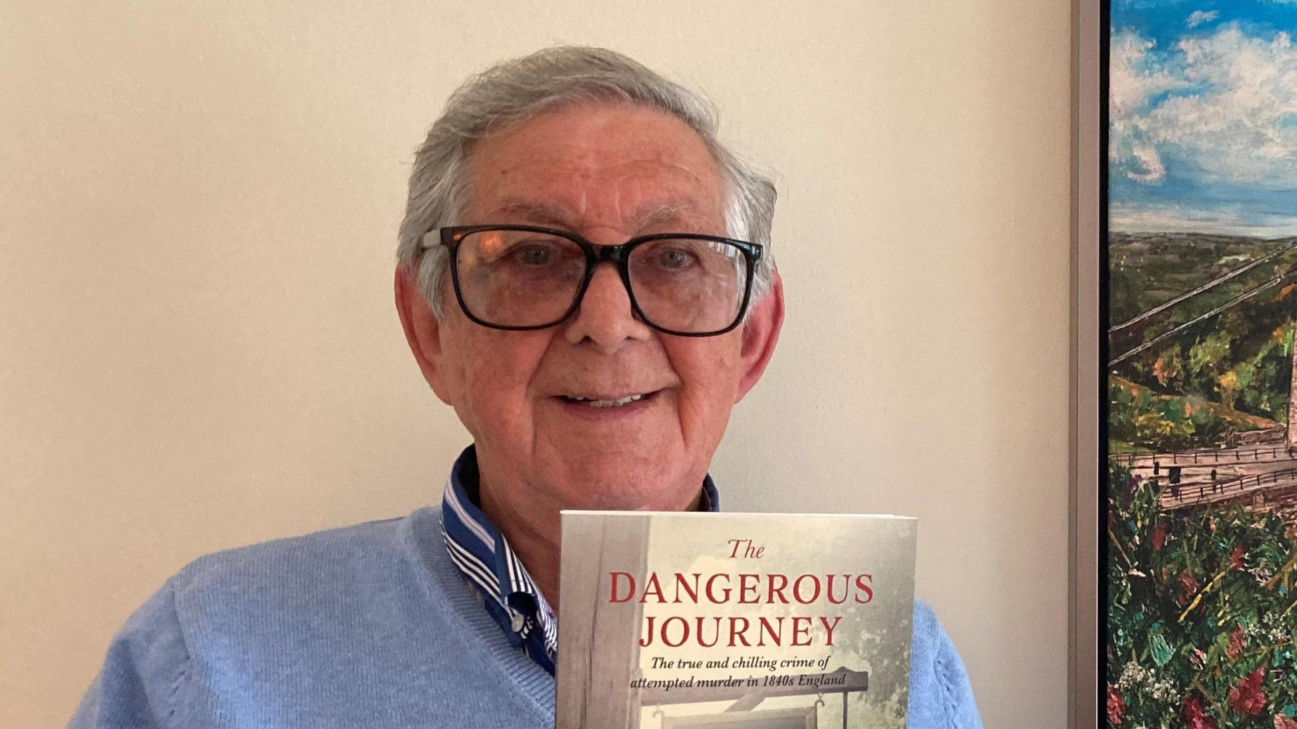 A man with grey hair and black-framed glasses holding a book with the title The Dangerous Journey written on it