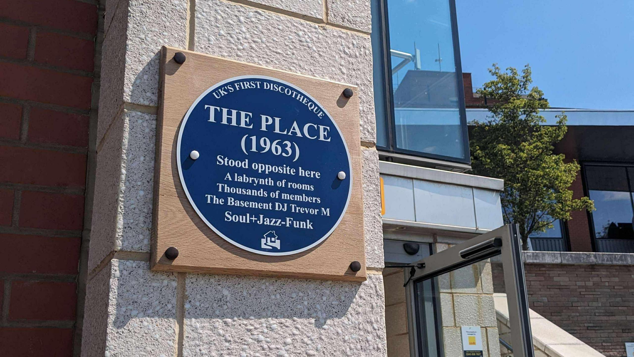 A blue plaque which is mounted onto a wooden square on the side of a building. The plaque has white writing which is titled "The Place" and details the site's history.
