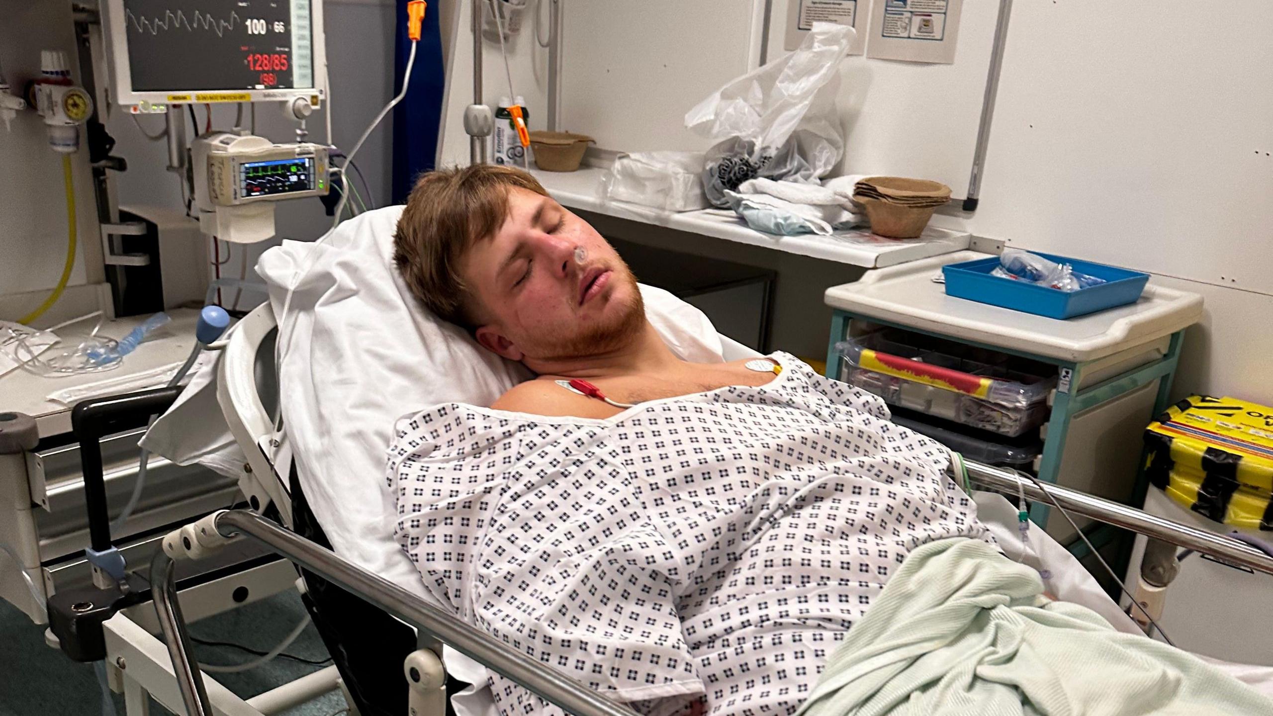 A man who appears asleep lying on a hospital stretcher wearing a white hospital gown and under a green blanket. Various items of medical equipment can be seen in the background.