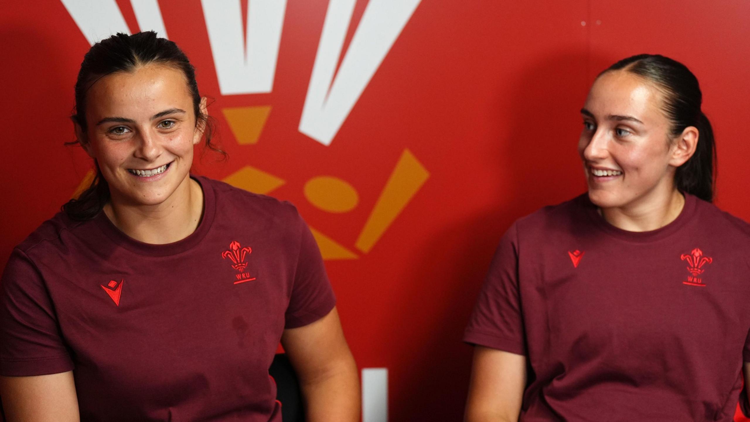Branwen faces the camera smiling, while Nel looks at her sister. They both have dark hair which is tied back, and are wearing burgundy shirts with the Welsh feathers on.  