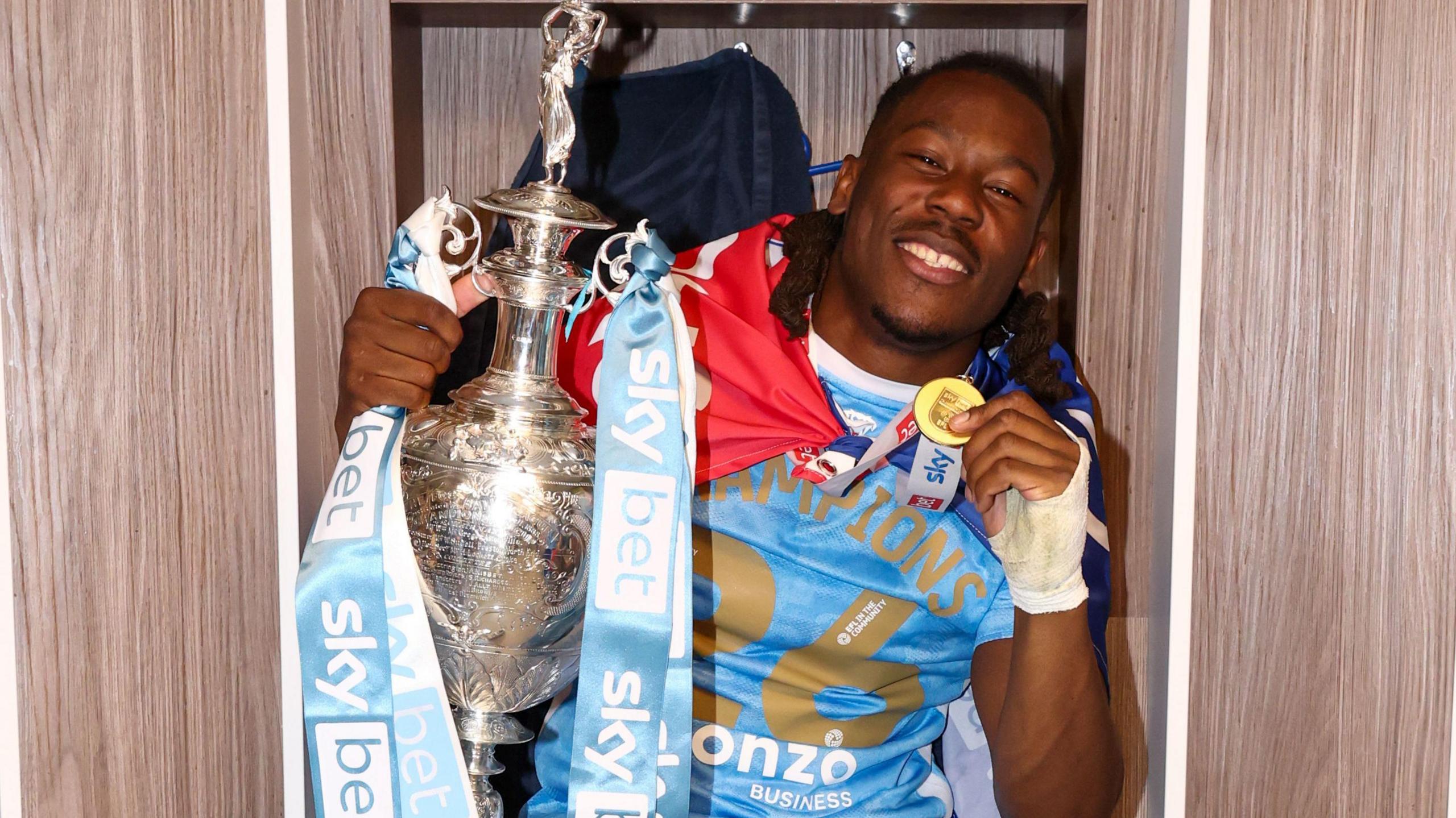 Brandon Thomas-Asante, wearing a sky blue Coventry shirt with 'Champions 26' written in gold on it, he holds the Championship trophy in one hand and his gold winners' medal in the other 