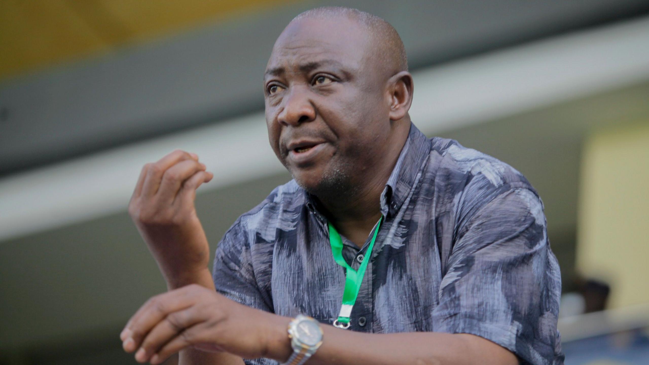 Ibrahim Gusau, wearing a black and grey shirt and with a green lanyard around his neck, gestures with his right hand as he stands during a football match. Gusau is visible from the stomach up as he leans forward on something which is just out of shot