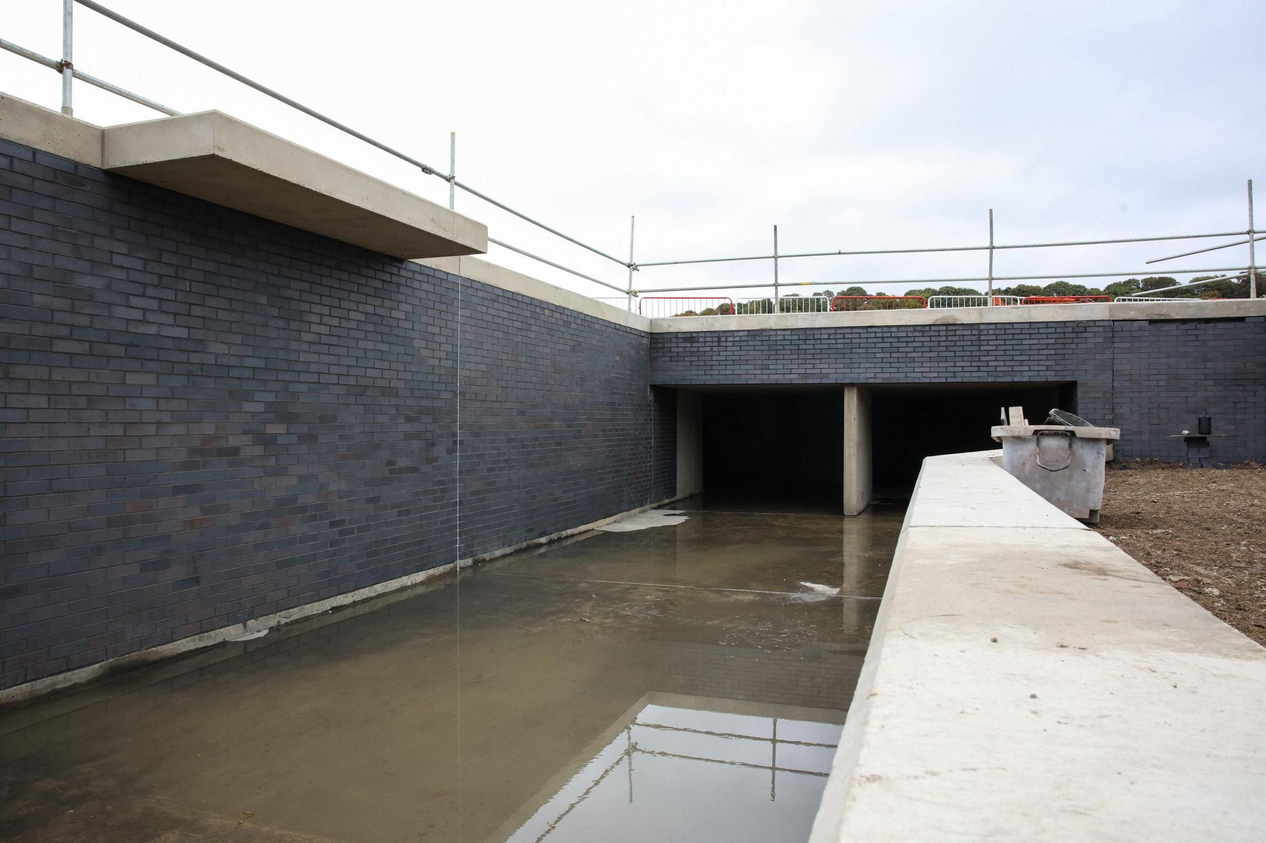 A section of the River Lea which runs between two walls, one high and one low. The walls are made of dark grey bricks, with a white concrete block on top. The water is very low level and murky. It then runs into a tunnel.