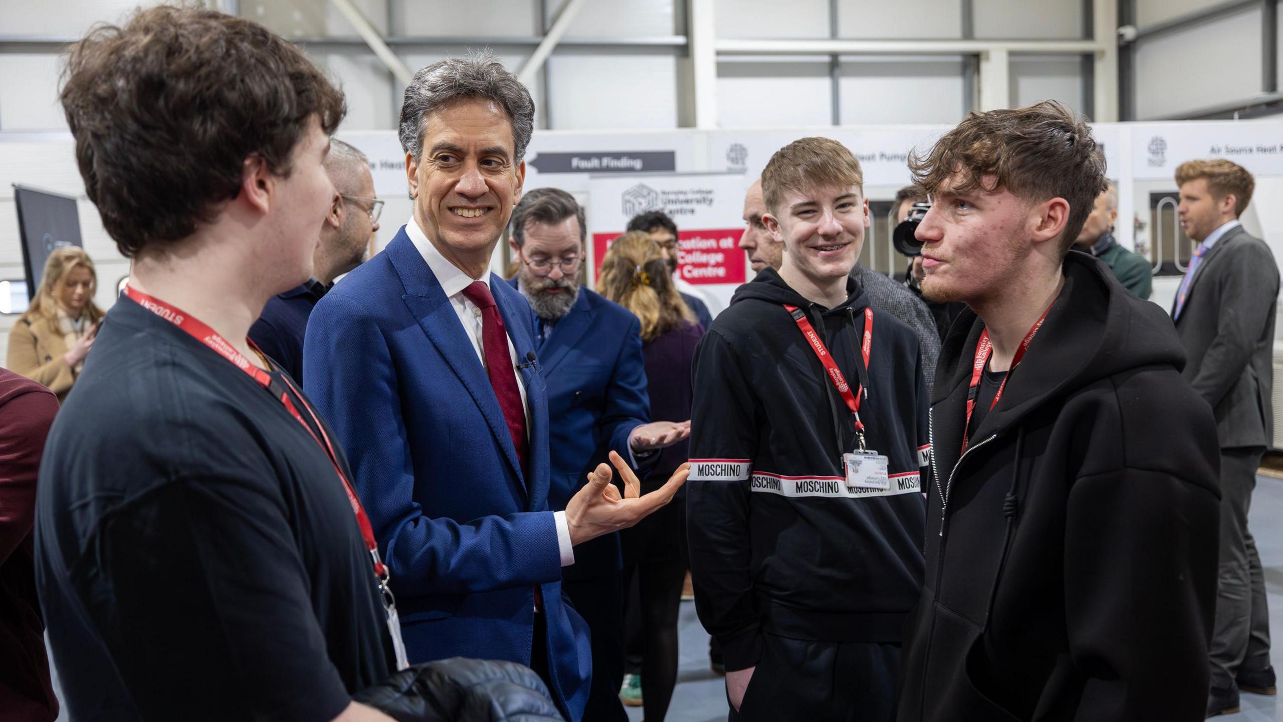 Ed Miliband, a man in a blue suit and red tie, meets students at Barnsley College to talk about jobs in the energy sector. The students are all male and are wearing black with red lanyards.