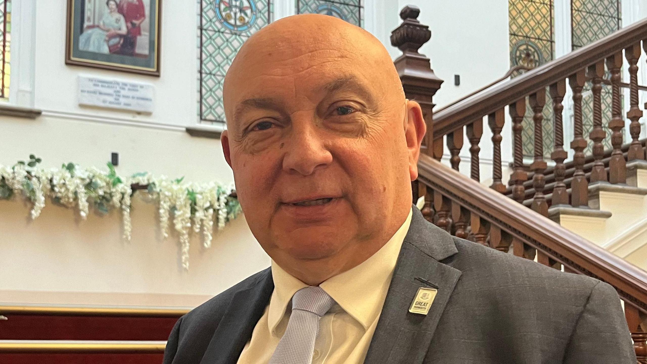 Carl Smith, a Conservative councillor who is standing inside a building on a staircase with red stairs. He is wearing a grey suit jacket, white shirt and light grey tie. He is looking directly at the camera and smiling.