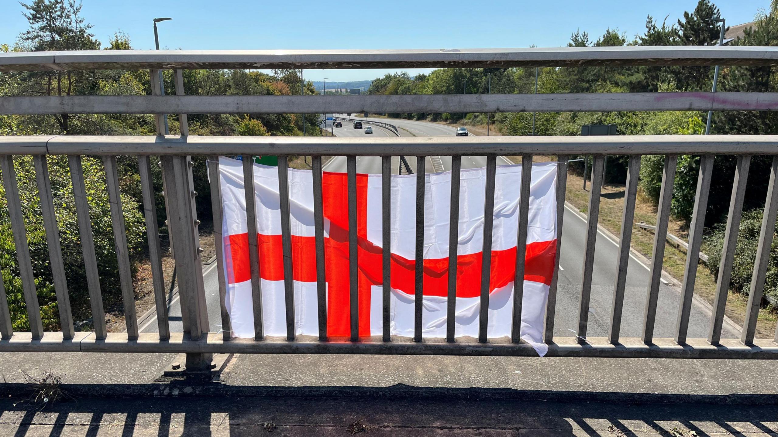 A St George's flag pinned to the railings on a bridge above a dual carriageway. The flag is on the outside of the bridge with the photo taking from behind it on the bridge. It is a sunny day with a clear blue sky and many trees lining the road.