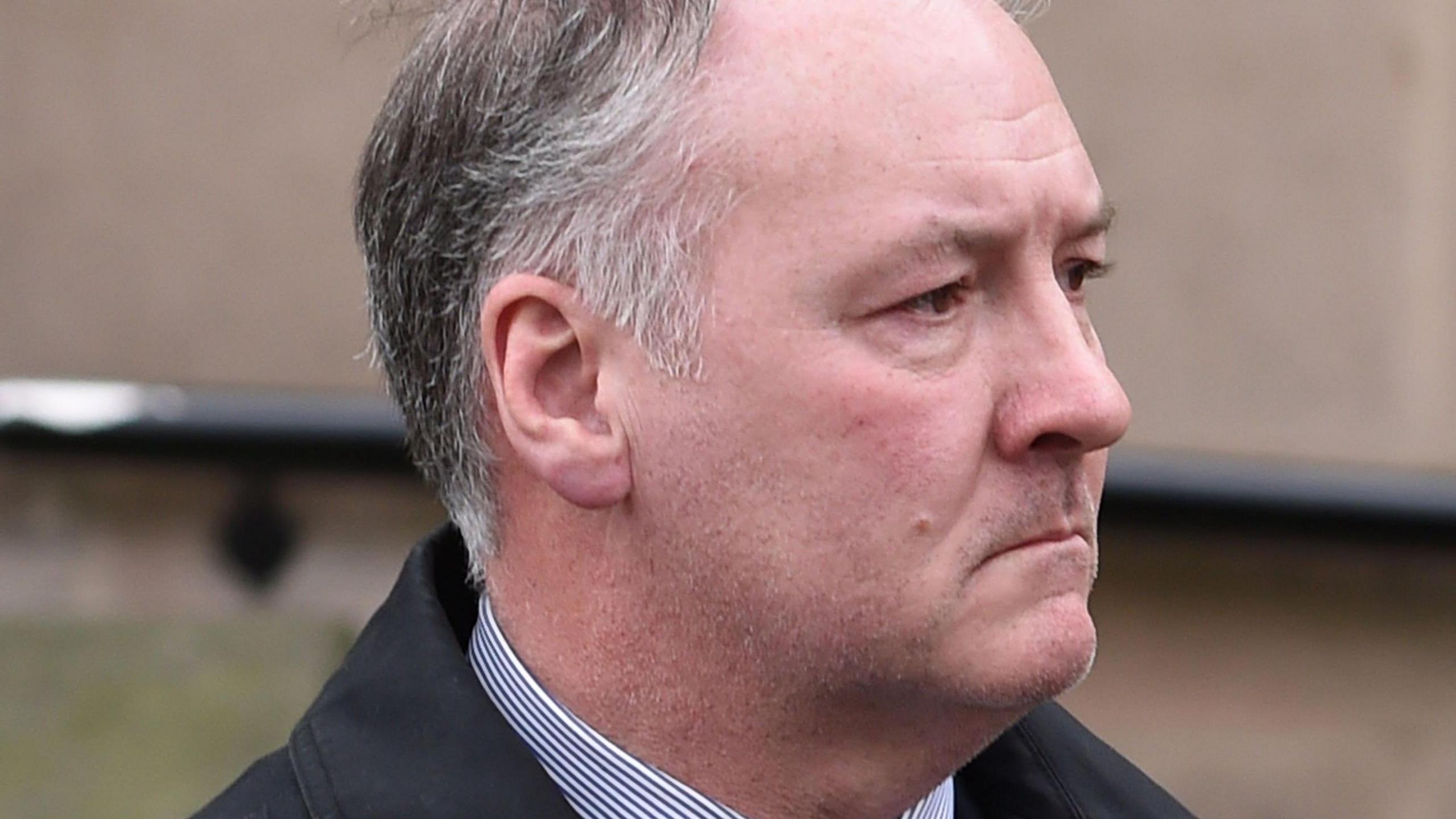 A close-up of a man's face from the side. A collar of a striped blue and white shirt and black jacket can be seen. The man has greying black hair cut in a short style, grey stubble and has a sombre expression on his face.