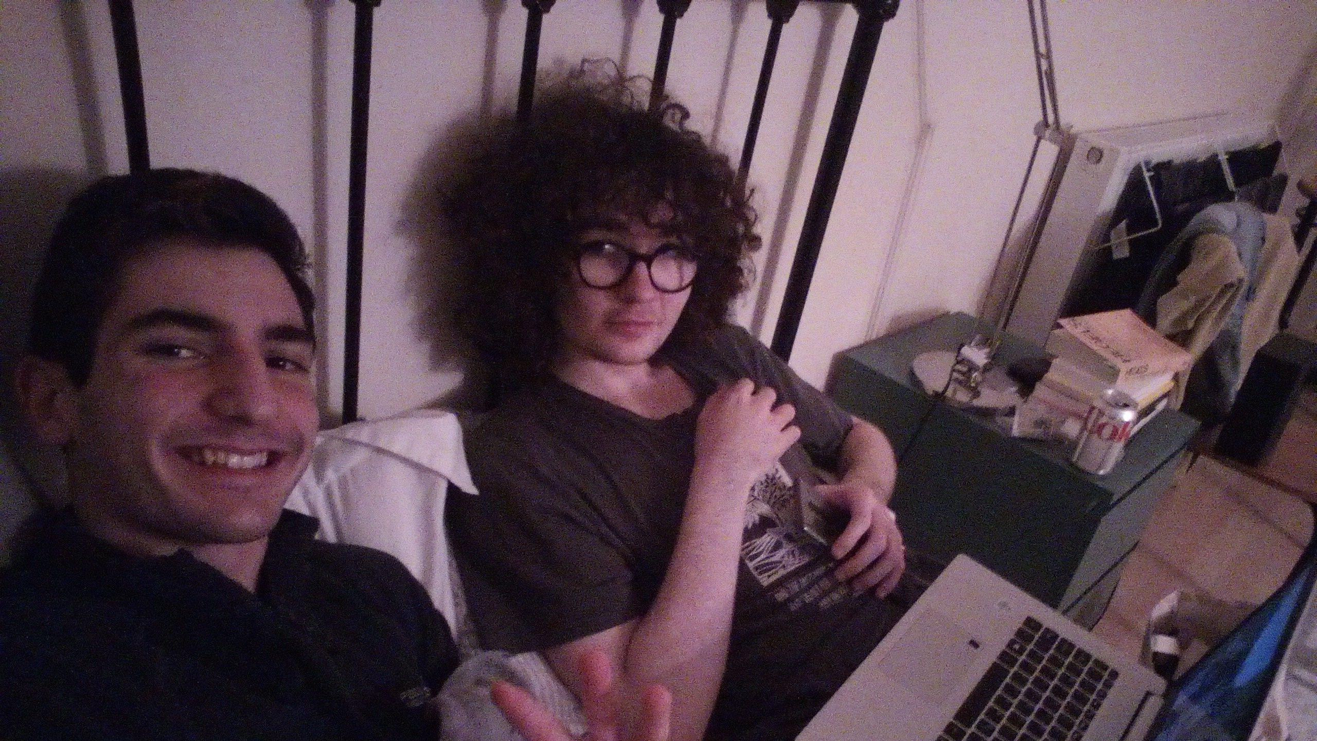 Two men sit on bed with a laptop. One man has dark curly hair and glasses and is holding a phone. The other has short, dark hair and is under the covers.