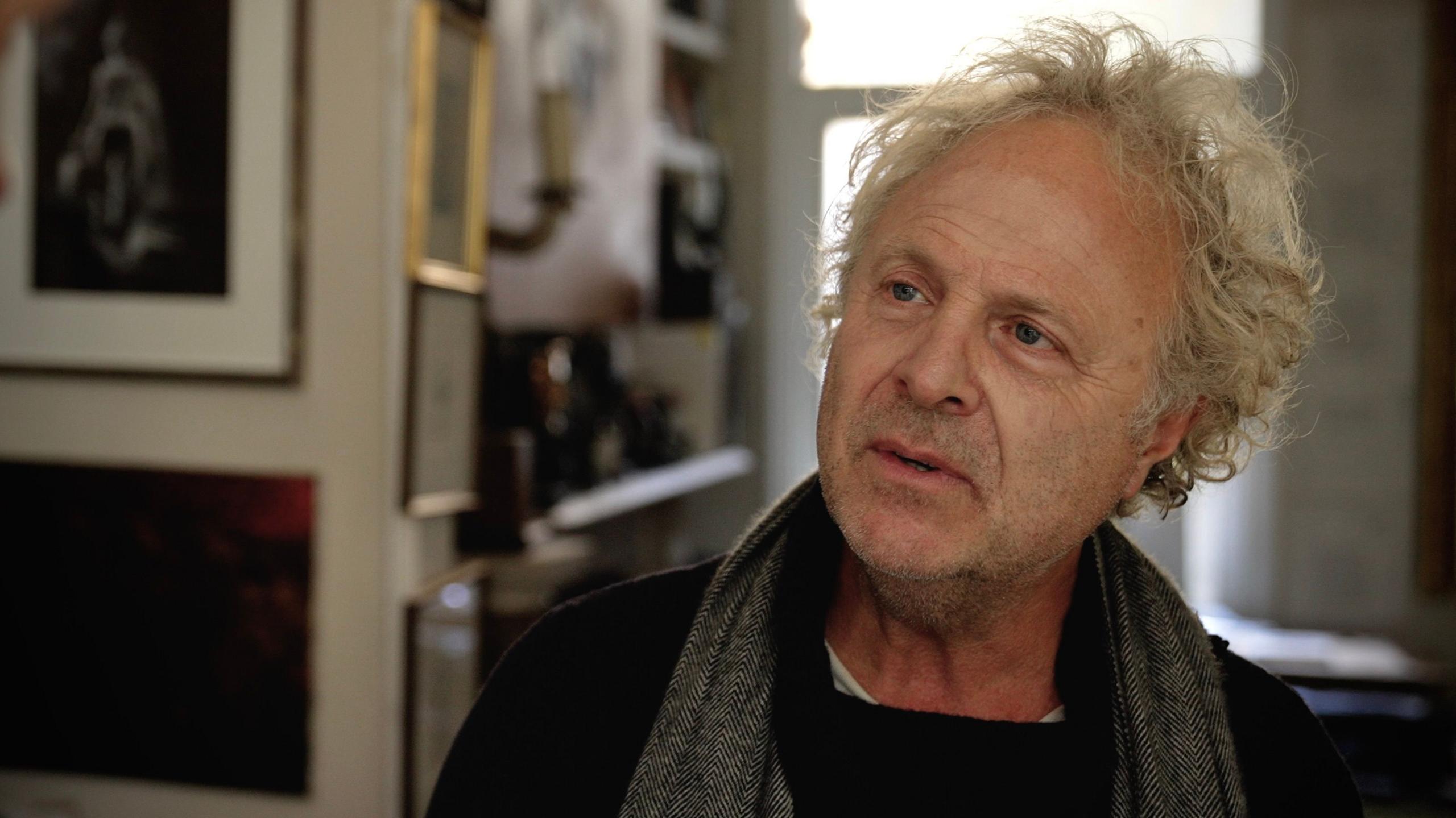 Charlie Mackesy with a scarf around his neck, in his home with pictures on the wall, he is highlighted by sun from the window behind him