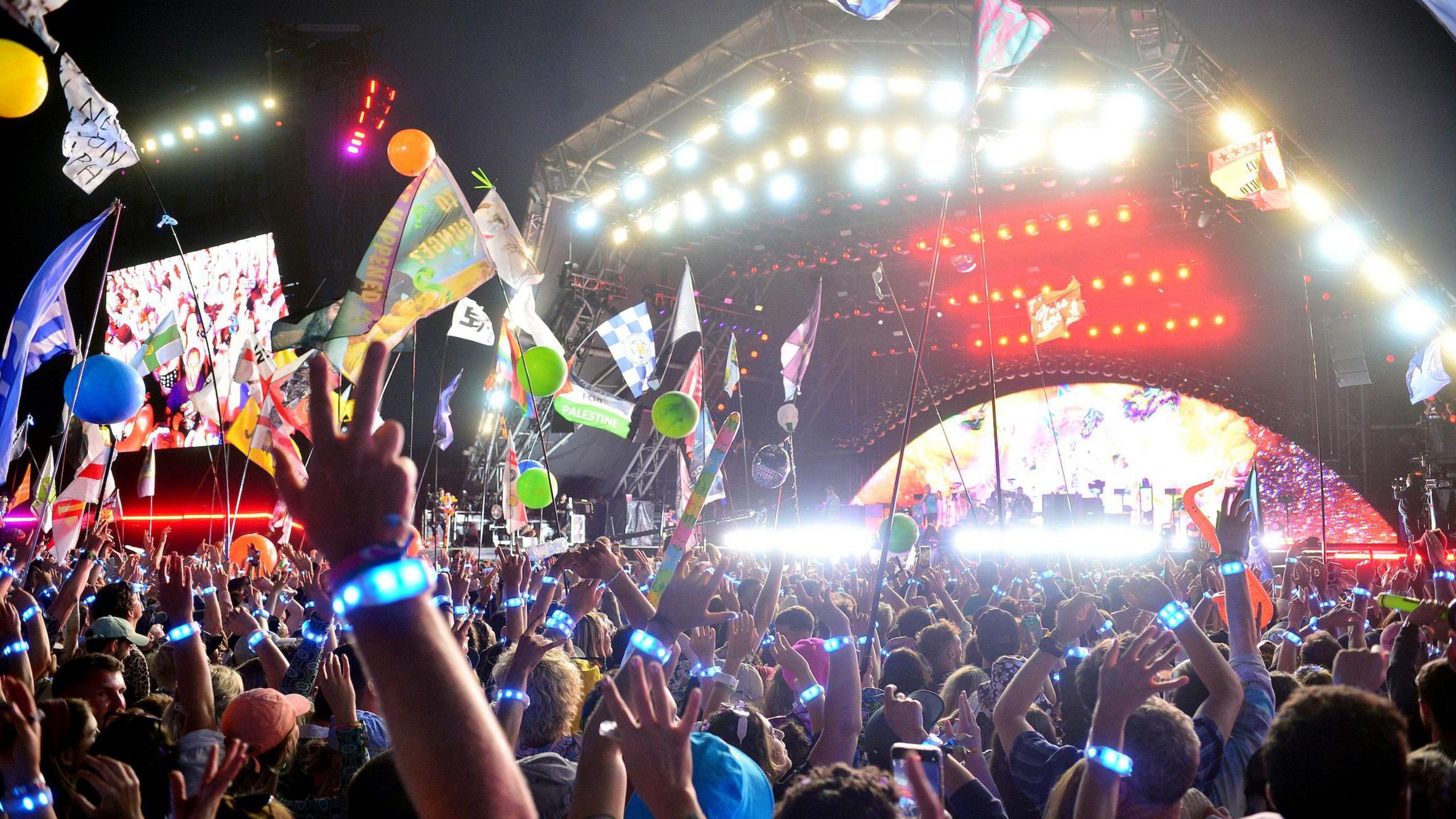Glastonbury Festival in 2024, showing the Pyramid Stage at night where Coldplay are performing. Bright lights are shining back at the crowd, who have their hands raised in the air, several waving flags, others making the peace sign with their fingers, and many wearing LED wristbands distributed by the band which give off a blue glow.