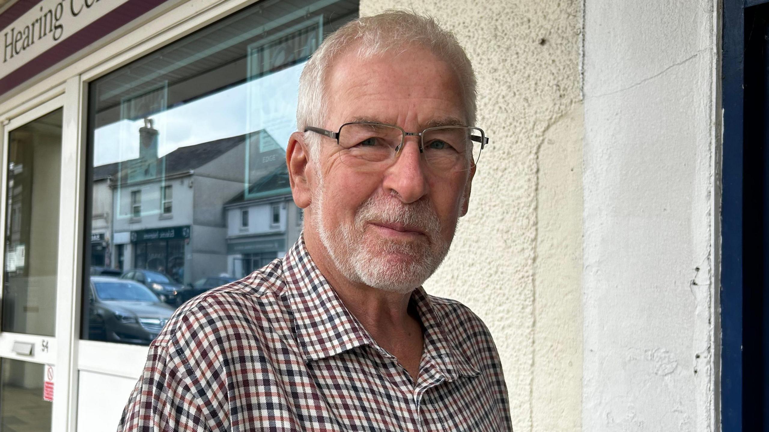A man, wearing a flannel shirt and glasses. He is looking at the camera with a slight smile. He is stood in front of a shop. There is a cream wall and windows and a white door. The logo on the shop says "the New Hearing Centre"