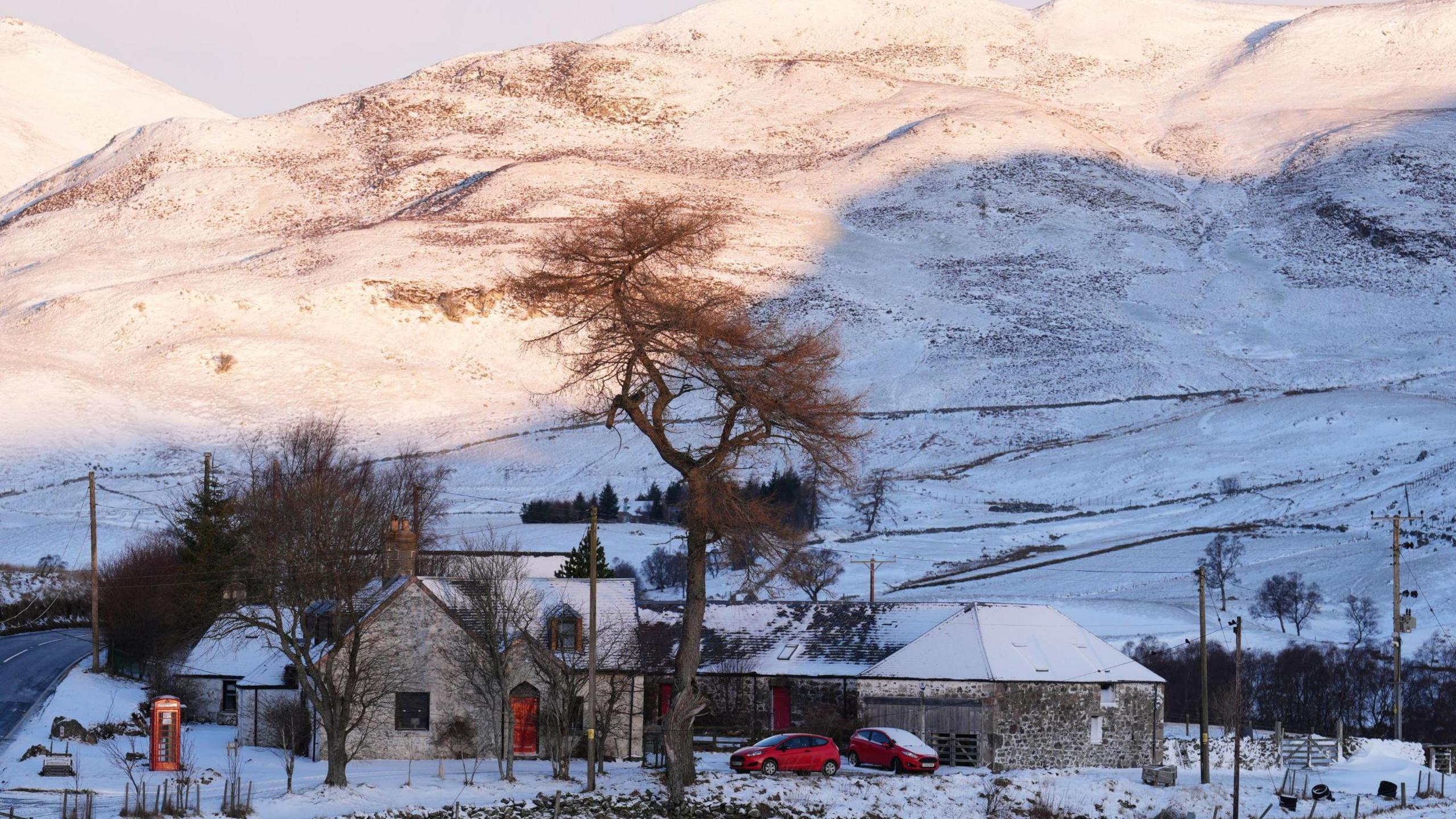 A couple of homes in Glenshee, with two red cars sitting in front of them. A red phone box is outside and hills are behind the homes. Snow covers the grounds. 