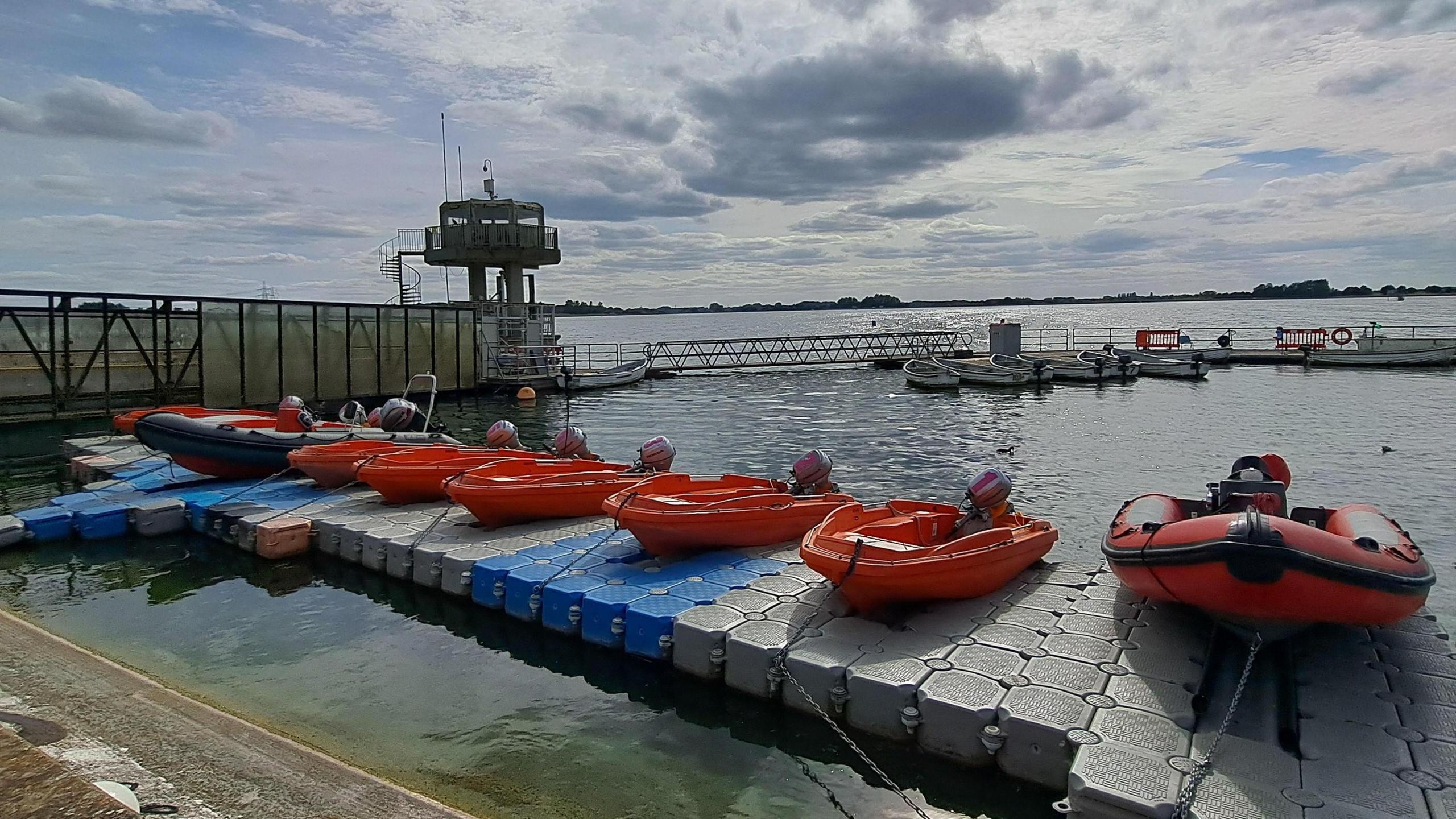  Several orange boats sit on a pontoon in the foreground. A cloudy bright sky with blue patches gives way to shimmering water of a reservoir