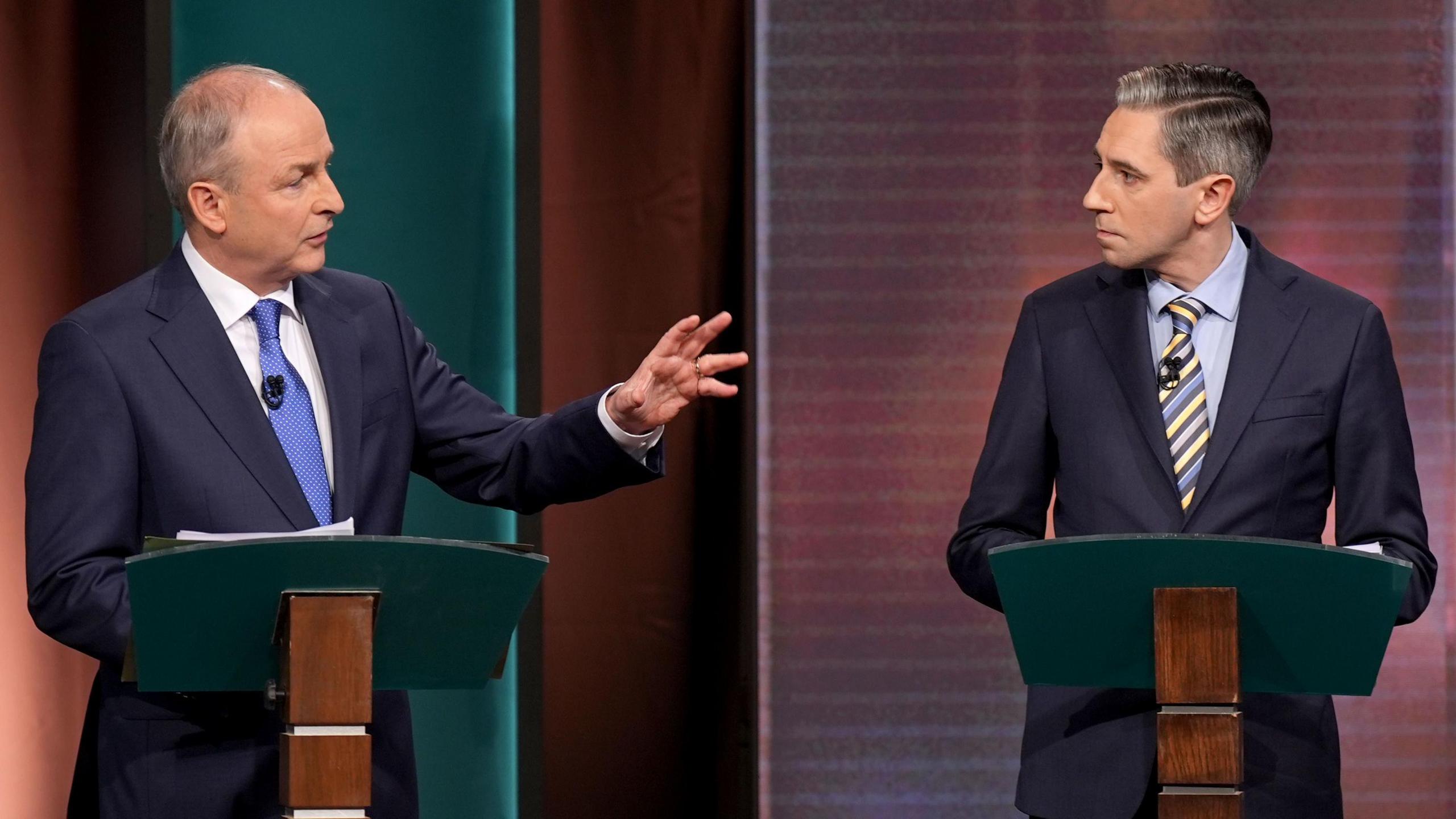 Micheál Martin and Simon Harris during the leaders debate. Micheal is wearing a dark blue suit, white shirt and blue tie - holding his hand up towards Simon. Simon is wearing a dark bule suit, white blue shirt and yellow and blue stripe tie. Both are looking towards the other and standing at podiums.