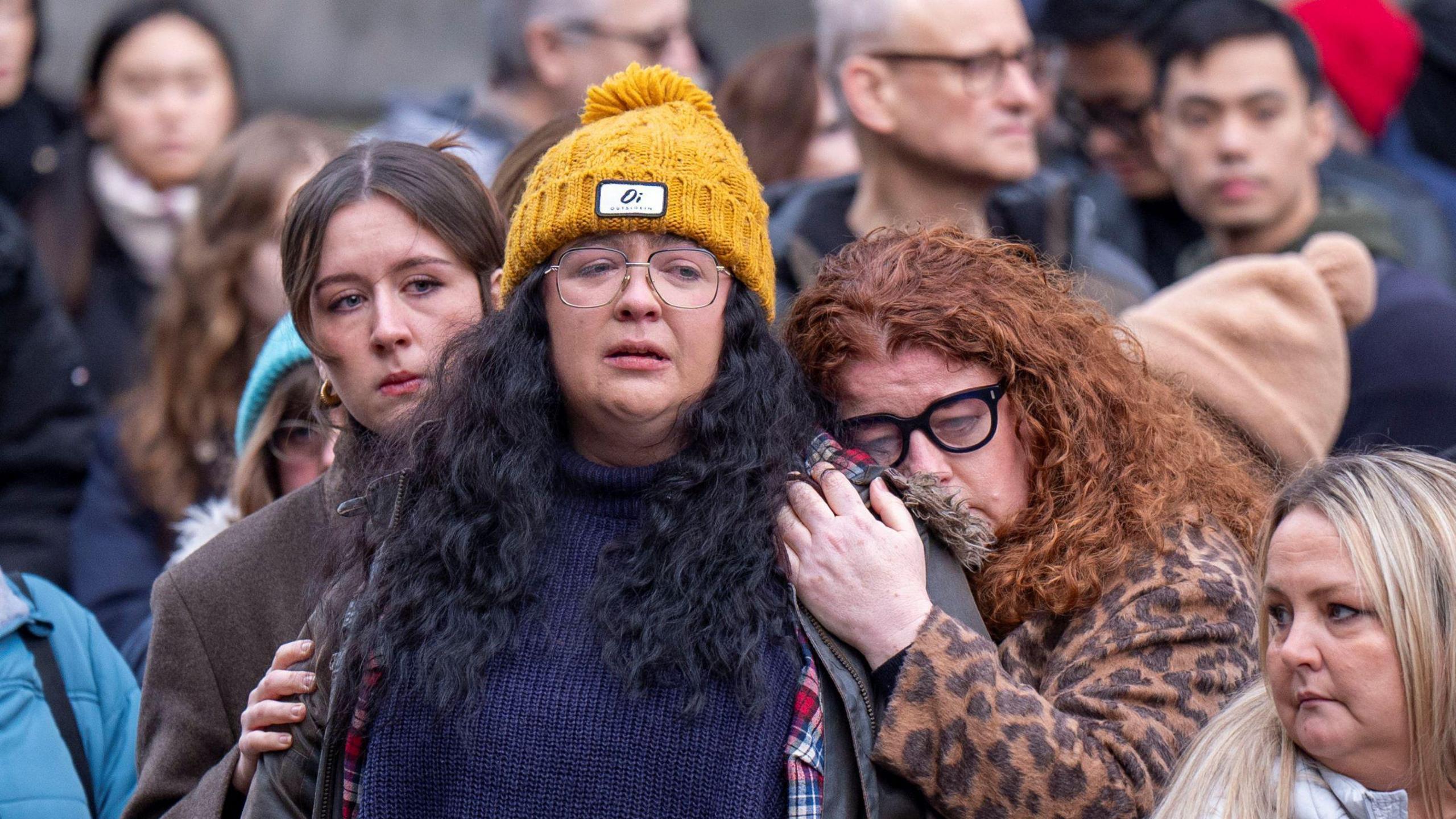 An emotional Ashley Storrie watching the hearse carrying her mum pass through Edinburgh. She is wearing a yellow hat and glasses and is being held by two other women, one of whom is red-headed and crying.