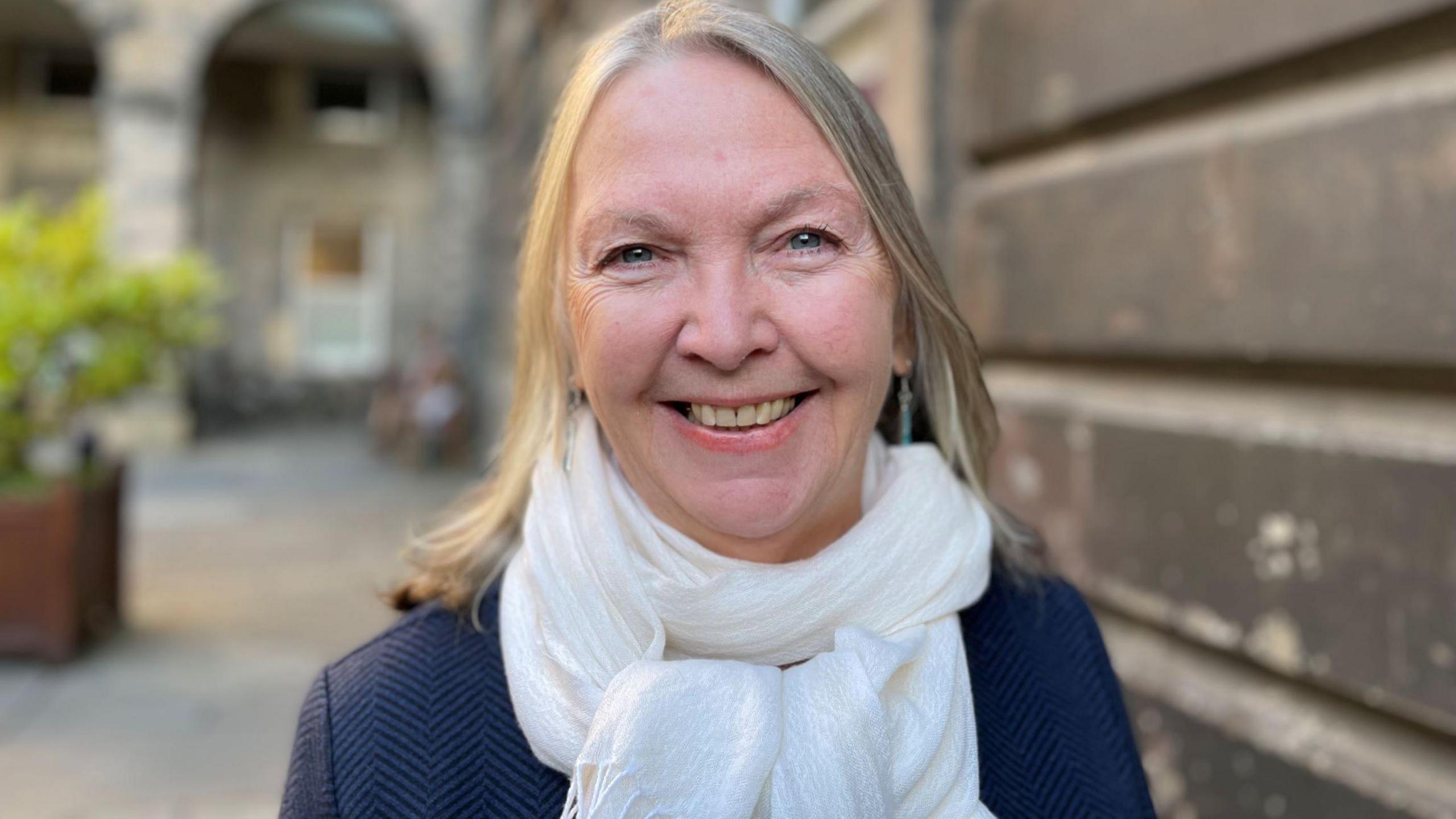 A smiling woman wearing a dark blue jacket and white scarf. She has blonde hair and is smiling directly towards the camera while outside.