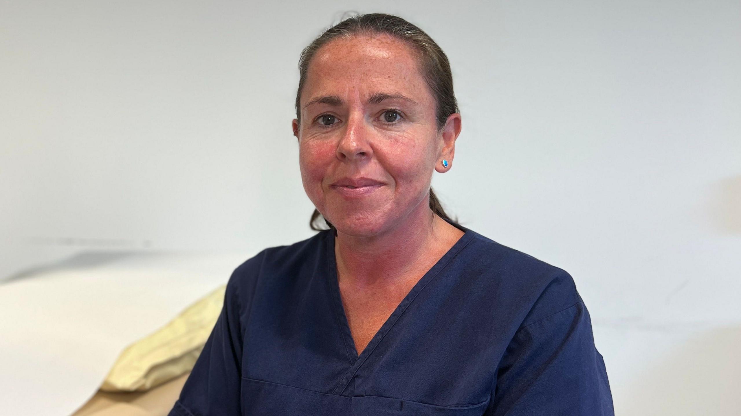 Corrinne Purdy in navy healthcare scrubs seated beside an examination bed in a clinical room. Medical equipment and a wall-mounted screen are visible in the background.