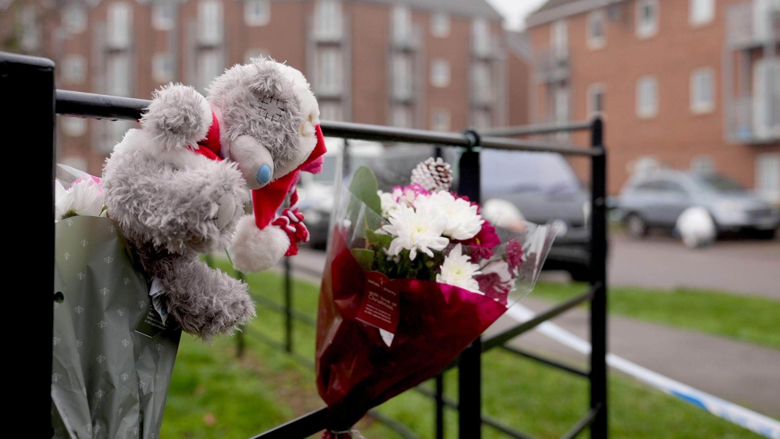 A grey teddy bear and two bouquets of flowers are attached to a black railing to the side of a residential road that is lined with blocks of flats. A police cordon tape can be seen blocking this road off from people.