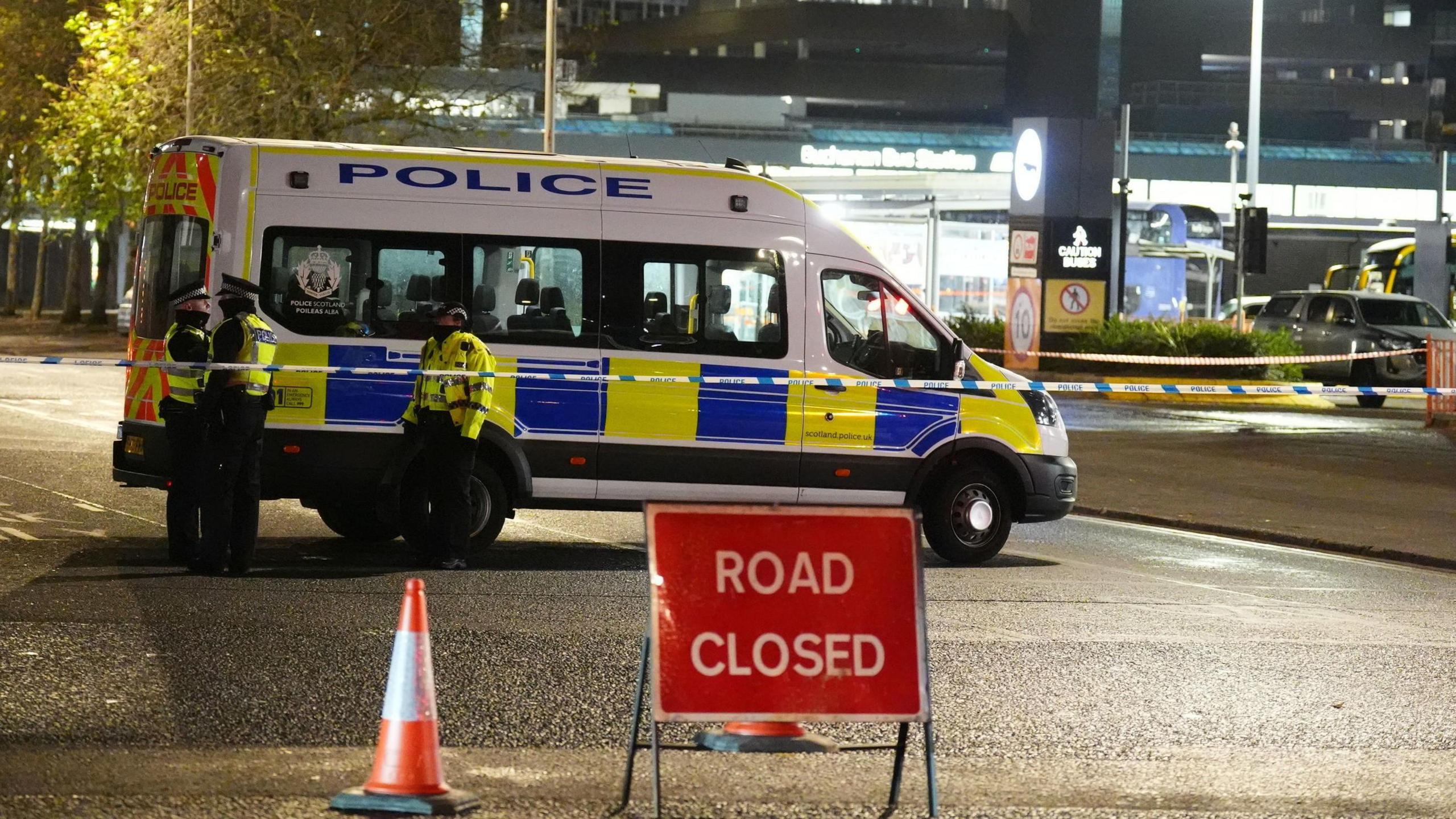 Three uniformed police officers stand next to a police van which is parked behind a road closed sign outside Buchanan Bush Station