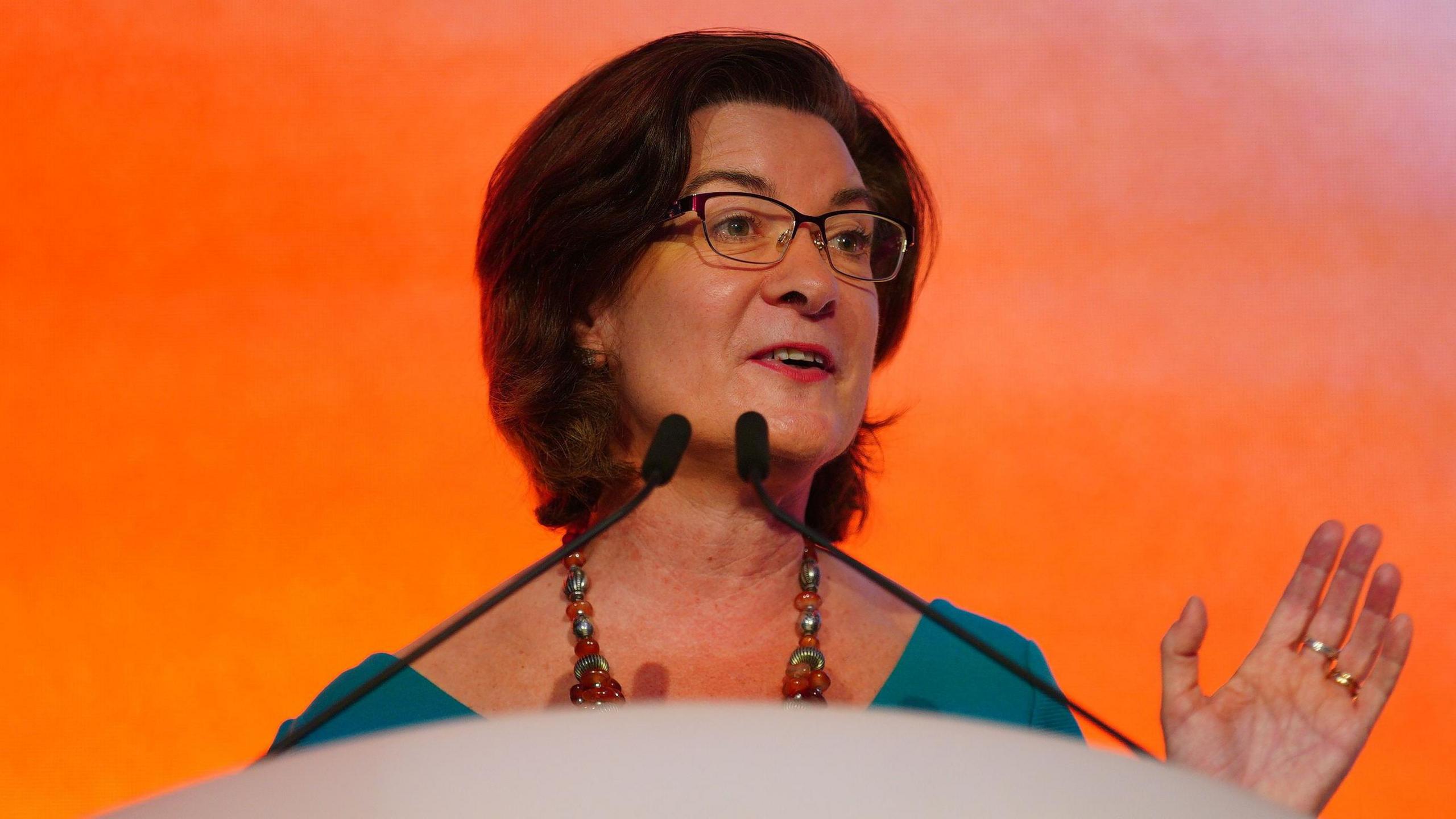 Eluned Morgan wearing a green top and a necklace; she is standing at two microphones, speaking, with an orange background