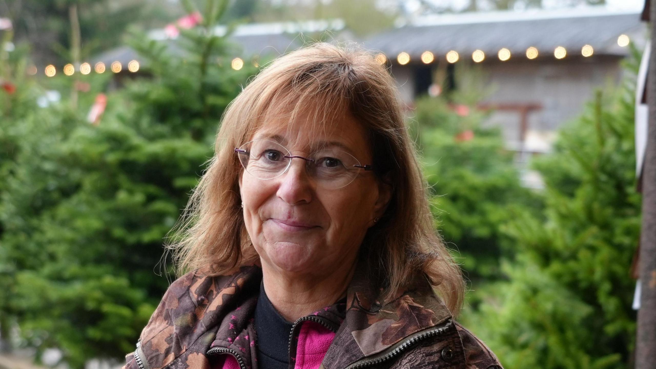 A woman with shoulder length brown hair smiles at the camera. She is wearing clear glasses with a brown camouflage coat. Christmas trees are pictured behind her. 