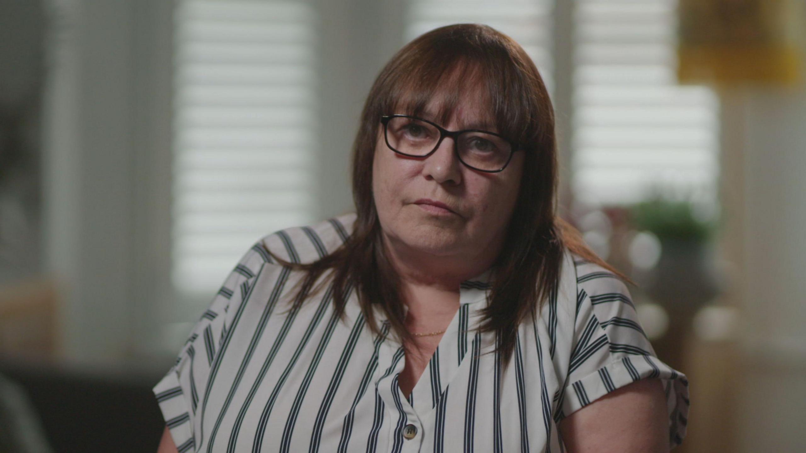 A woman with dark hair and thick rimmed glasses is sat in front of a blurred out living room, which has a plant and shutter blinds in the background. She is wearing a cream and black pin-stripe shirt and looks towards the camera.