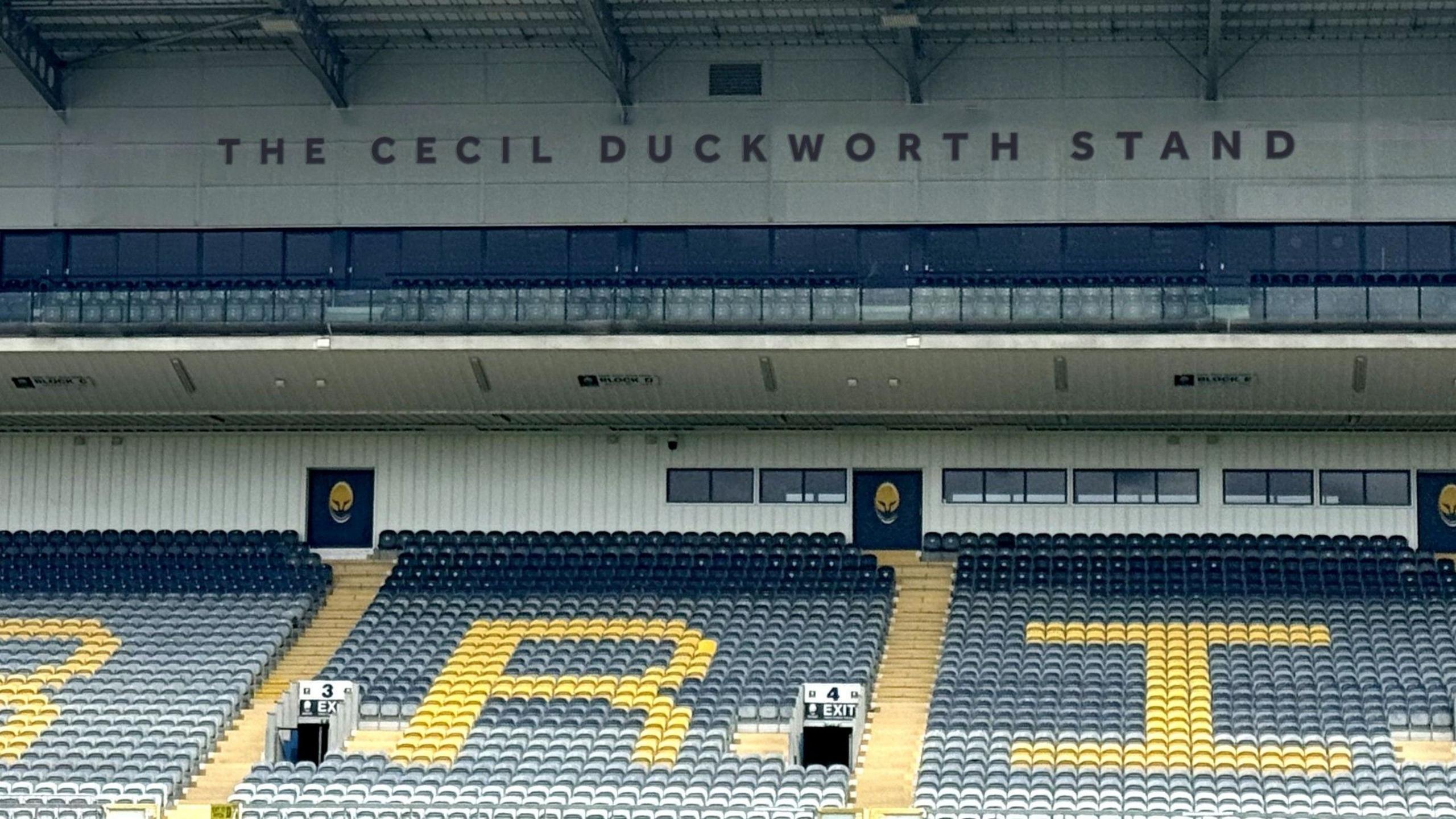 The stand as viewed from the pitch's halfway mark. A large single-tier section of blue seats are shown, with interspersed yellow seats spelling out some of the letters of Warriors - the letters R and I are depicted. Above that, under the stadium roof is the wording The Cecil Duckworth Stand. The wording is in blue, on a white background, and in capital letters.