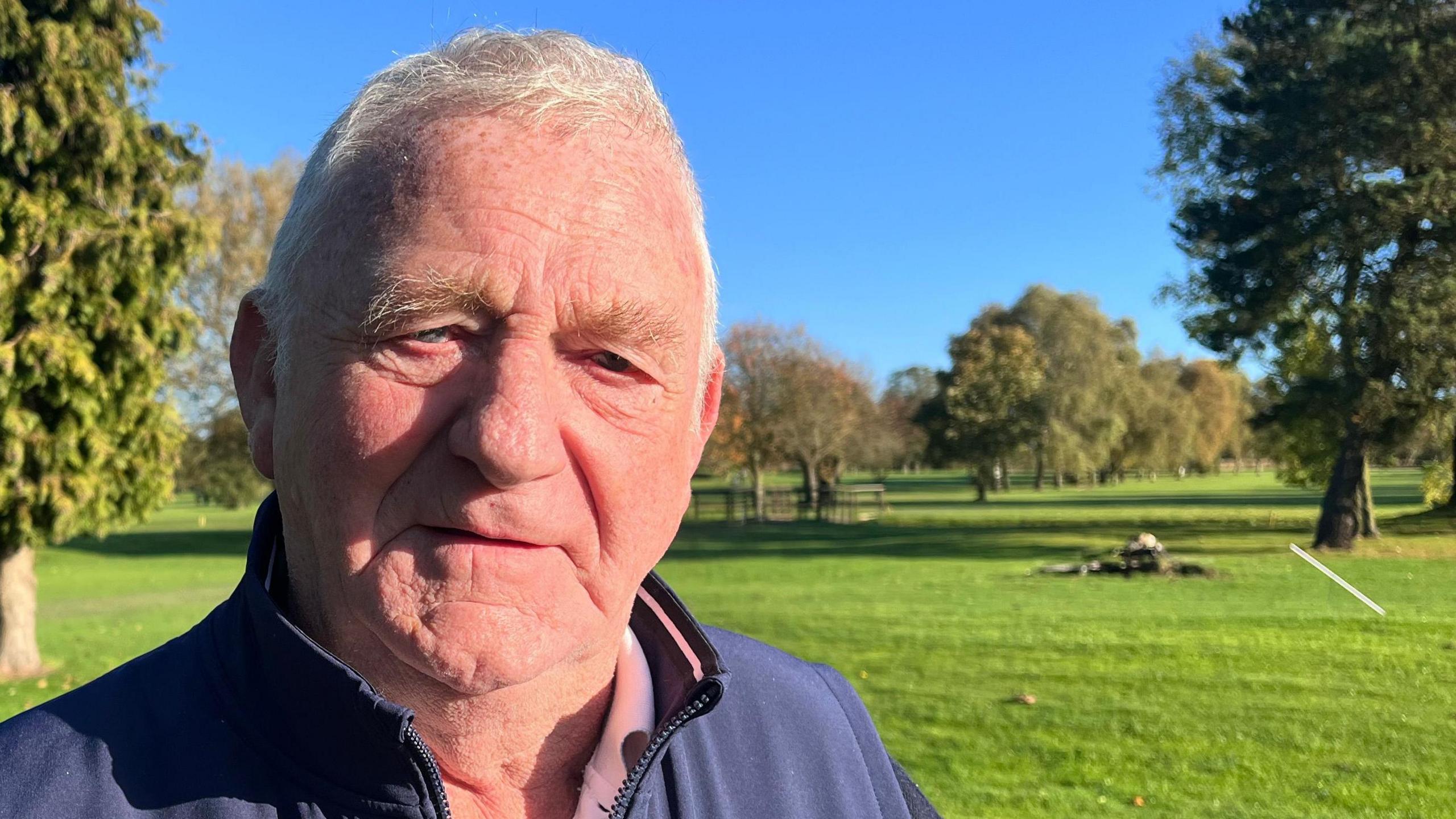 David Thomson stands beneath a blue sky on a green fairway at Ganstead Park golf club. He has short, white hair and wears a light-purple fleece.  