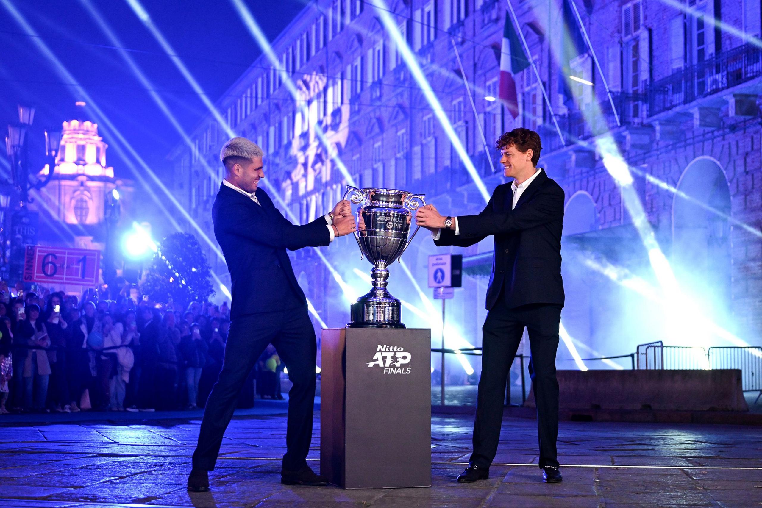 Carlos Alcaraz and Jannik Sinner in formal suits holding a large silver trophy on a pedestal labeled ‘Nitto ATP Finals,’ with bright blue and white spotlights illuminating a historic building and a crowd in the background during an outdoor evening event