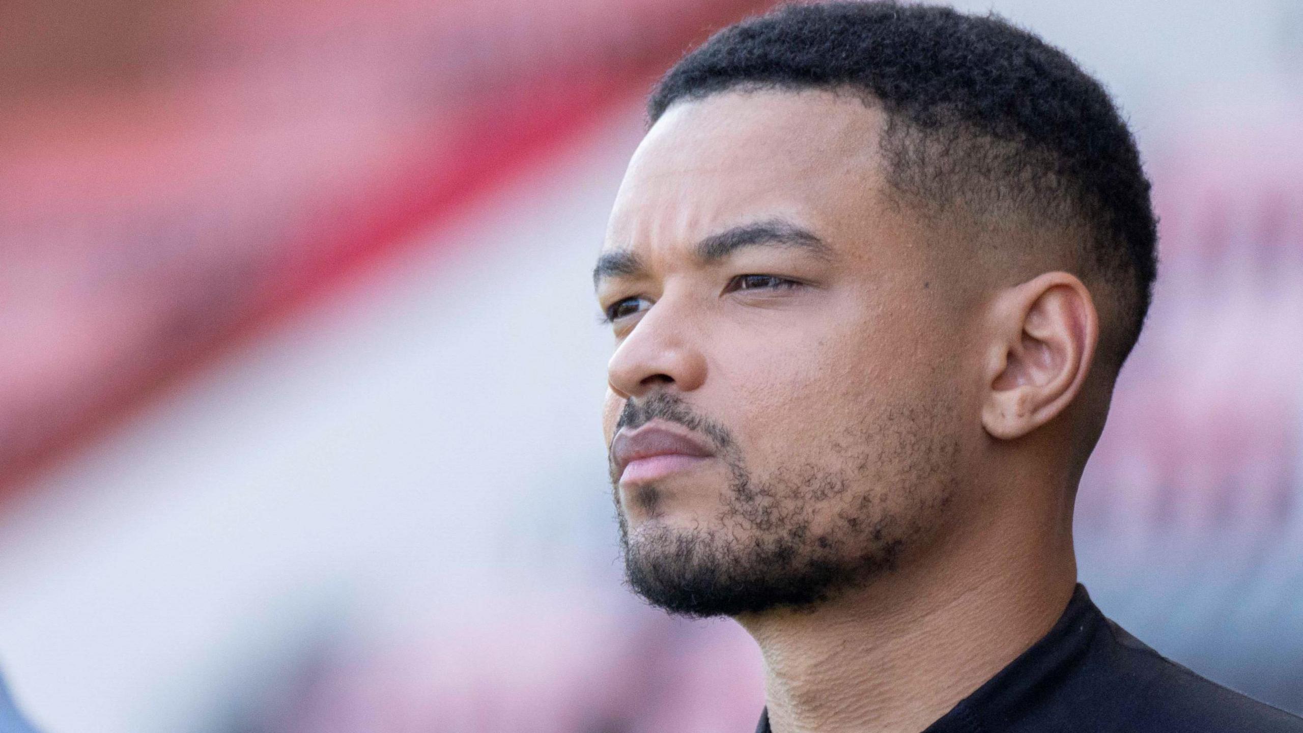 Steven Bartlett, pictured with a serious expression at a football match. Bartlett has short hair, a short beard and brown eyes. He wears a black T-shirt. 