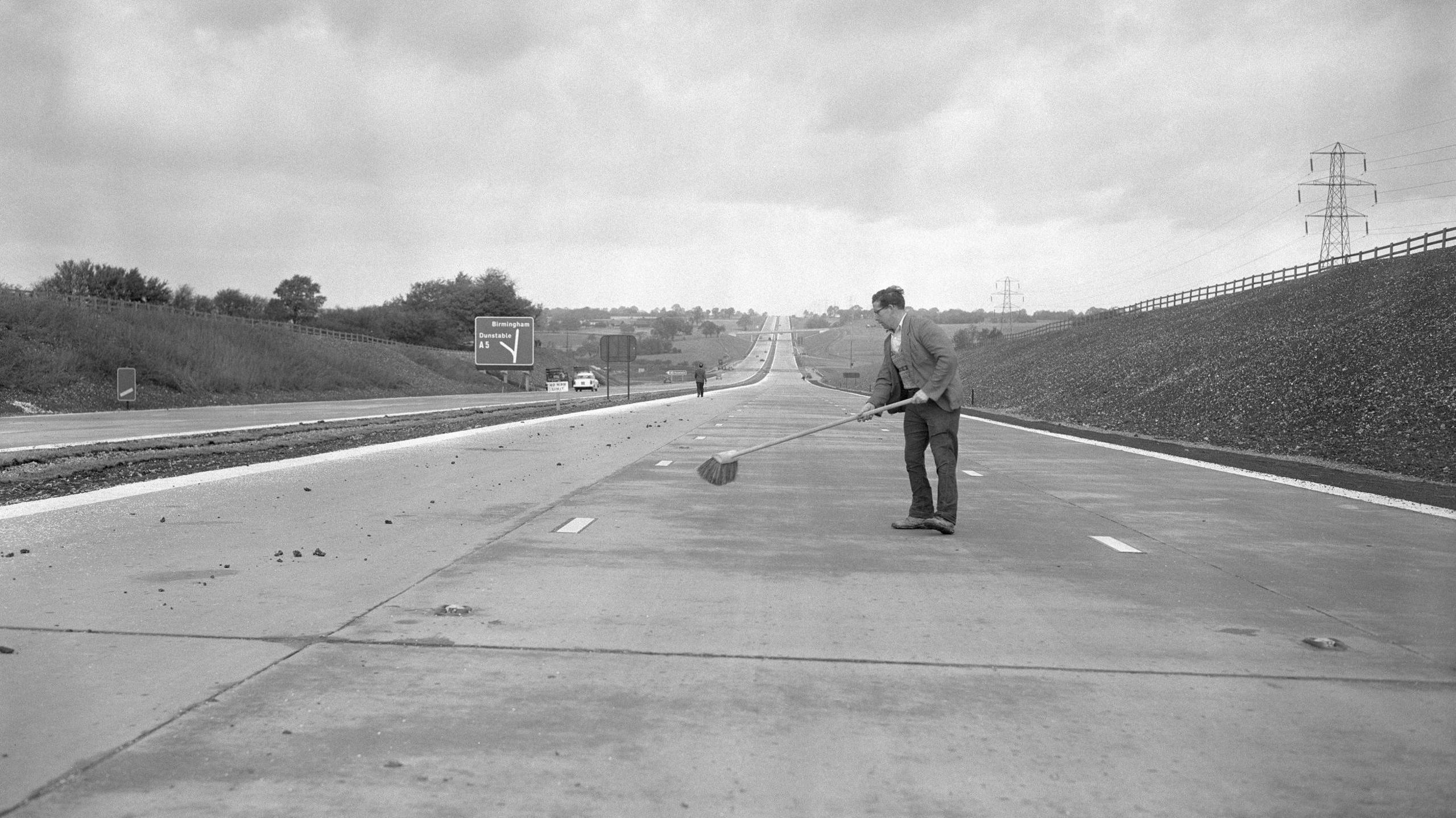 A black and white photo of a man holding a brush cleaning part of a newly construction motorway, dressed in an unexpectedly smart jacket