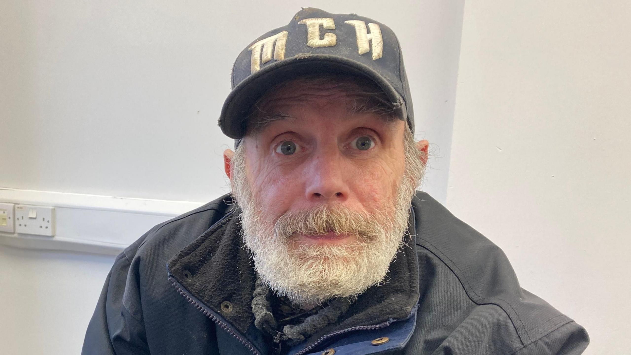 Gary Morgan, wearing a black jacket and a blue faded denim cap, looks into the camera. The cap has the letters M-C-H on it. He is sat in a room with white walls