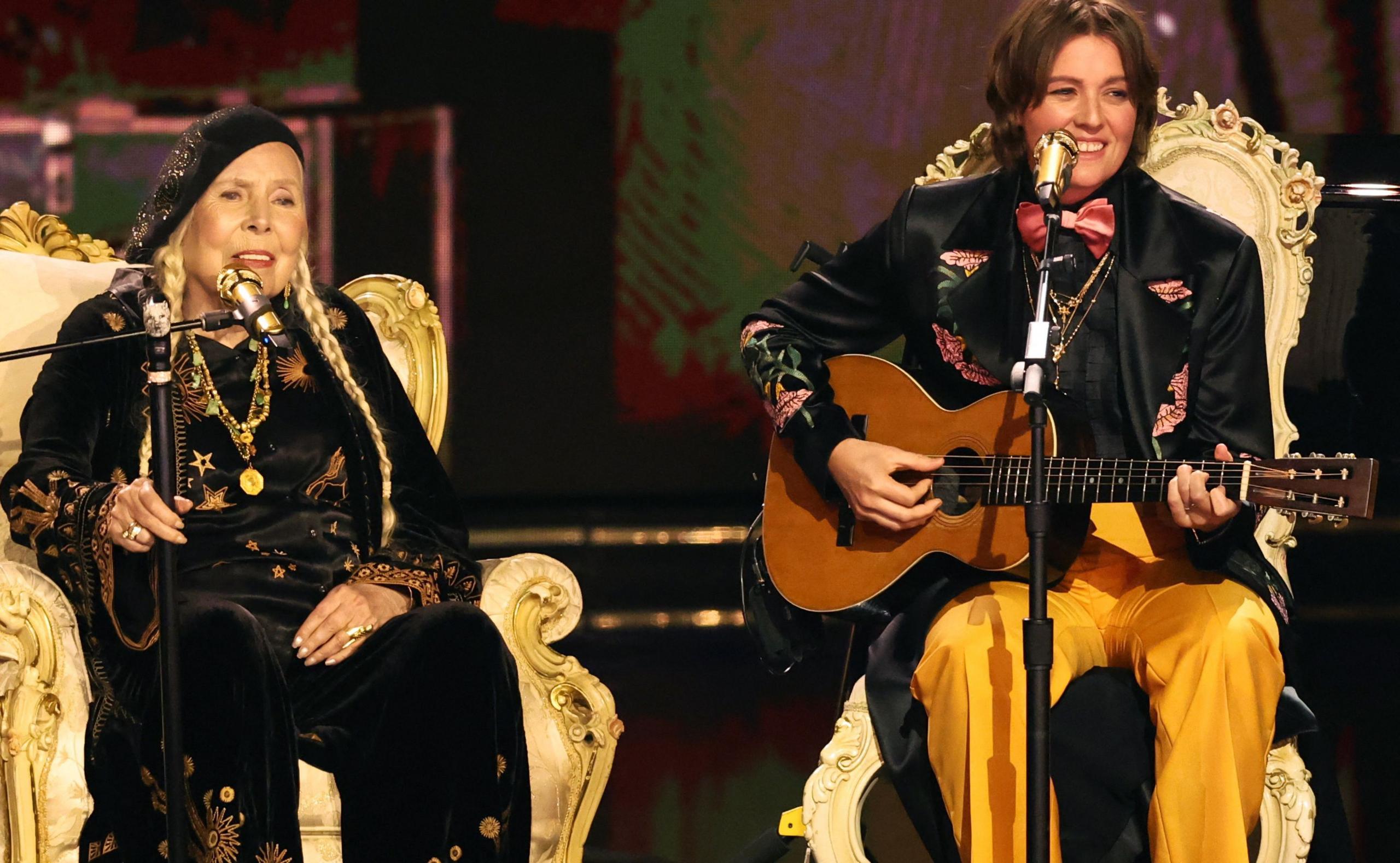 Joni Mitchell on stage with Brandi Carlile. Both musicians are sitting on thrones and singing into golden microphones