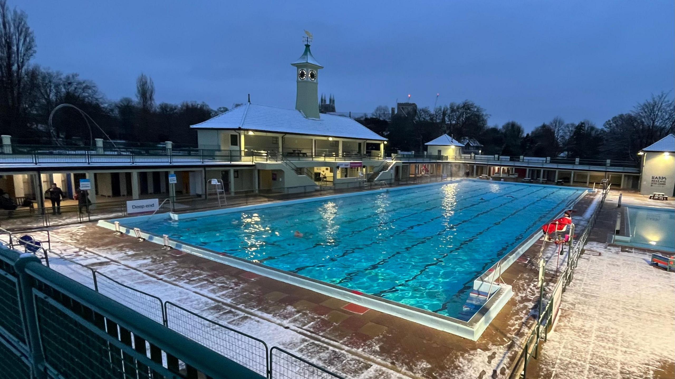 An outdoor swimming pool at twilight, with clear blue water and snow sprinkled around its perimeter