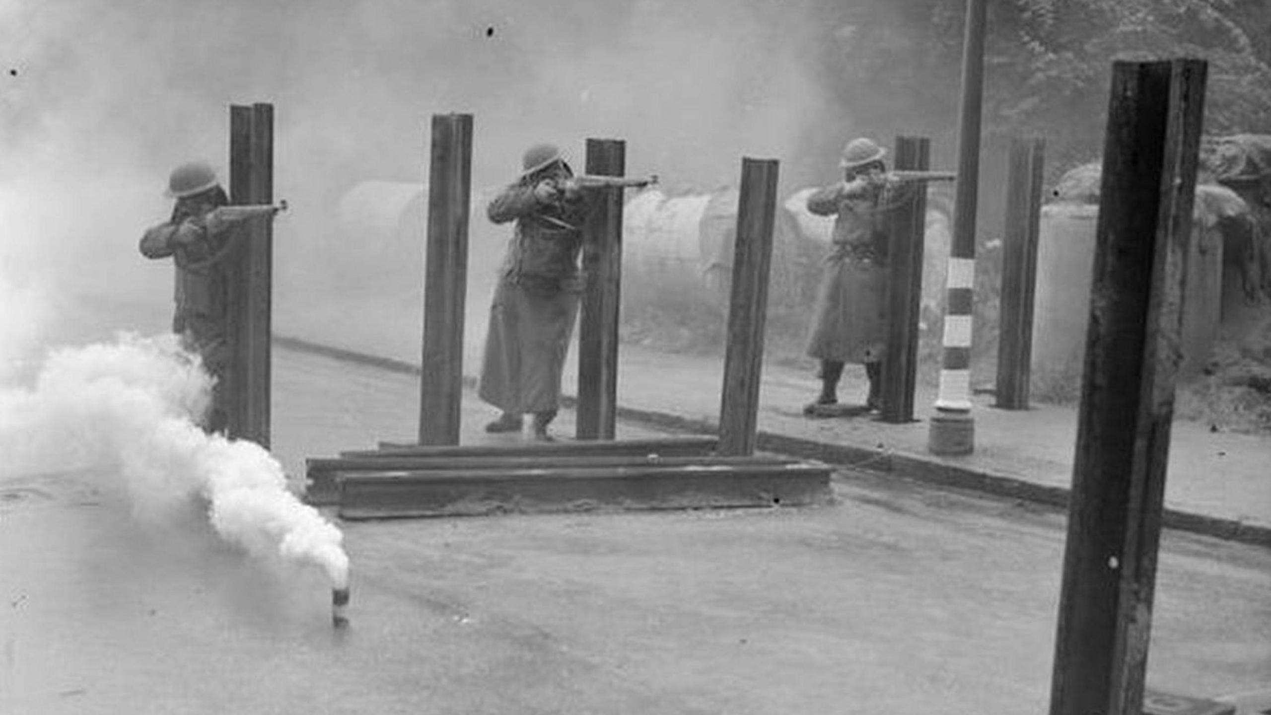 Black and white photograph of the three soldiers from the Home Guard. They are firing rifles with a smoke bomb in the foreground. 